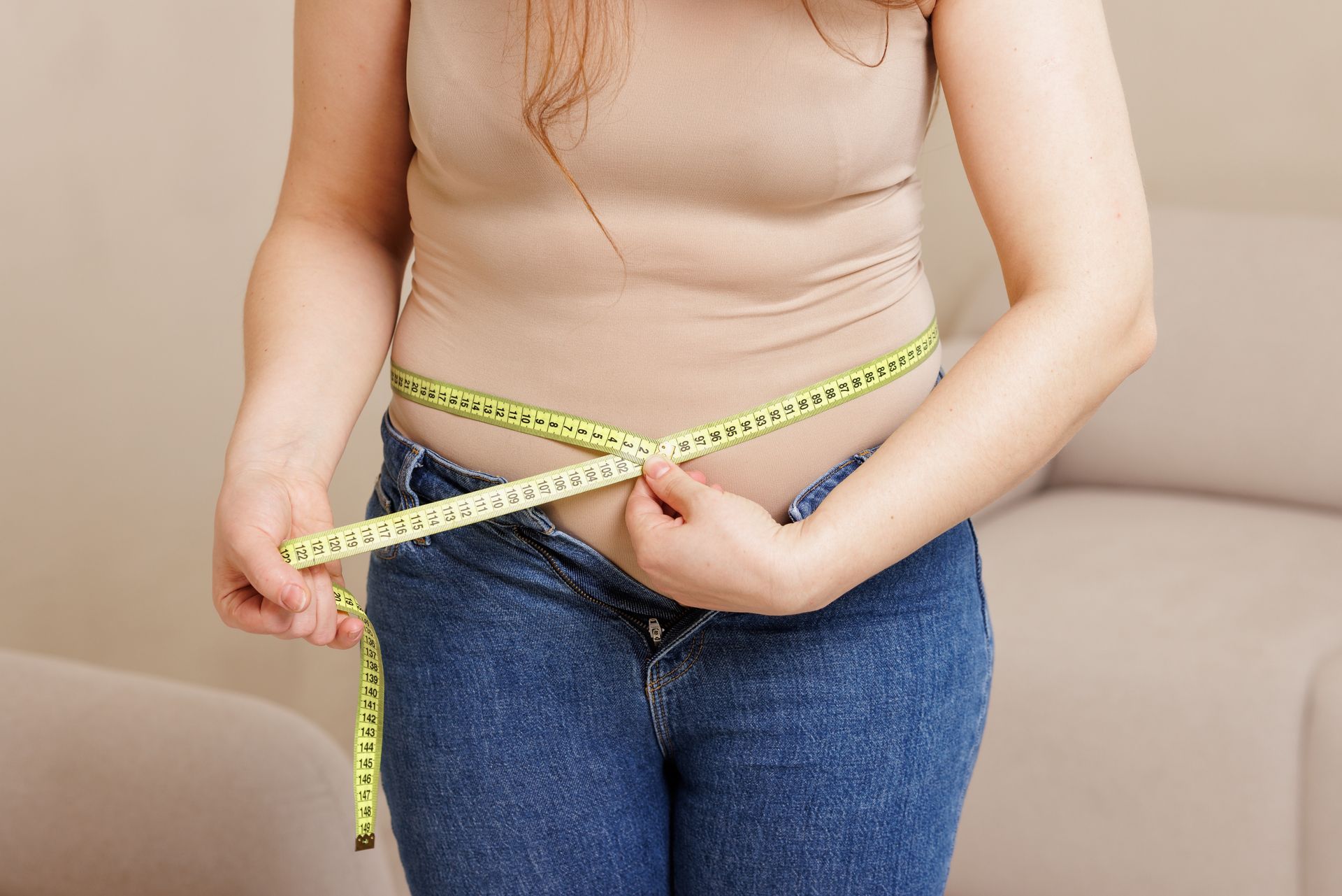 Person in beige top and jeans measures waist with yellow tape measure.