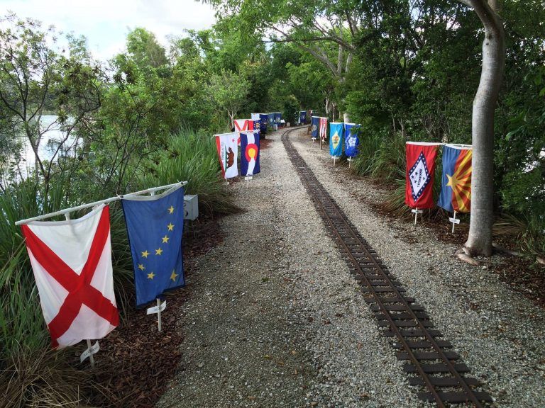 A gravel path with miniature train tracks flanked by various flags, leading through a wooded area near a lake.