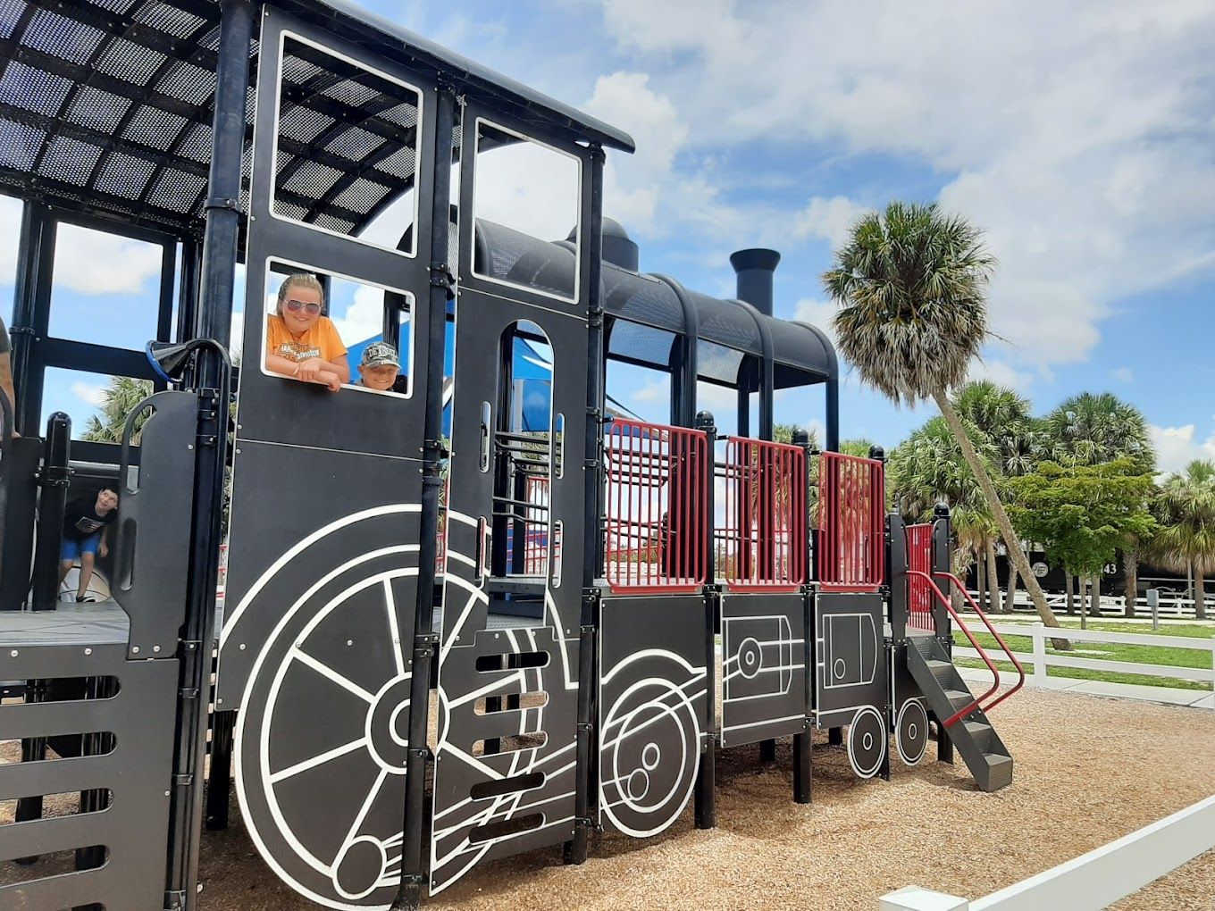 Playground with a black train-shaped structure; two children look out of the train windows under a cloudy sky.