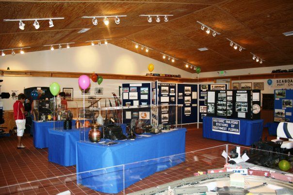 An exhibit hall featuring display tables with historical items and informational boards, adorned with balloons and overhead lighting.
