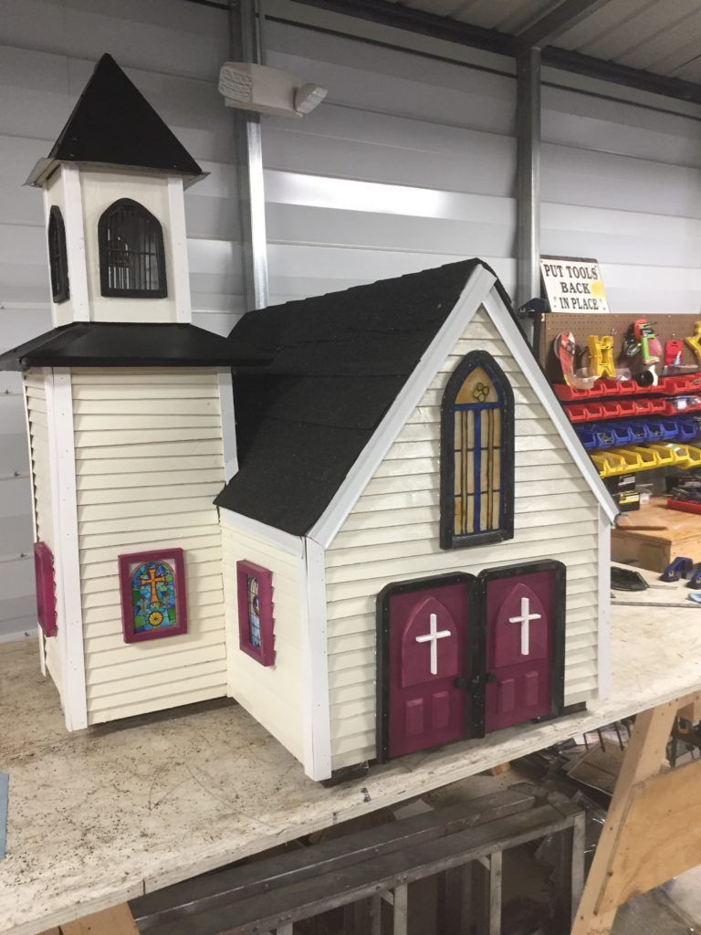 A miniature church with a white exterior, black roof, and purple doors and accents, sitting on a workbench.
