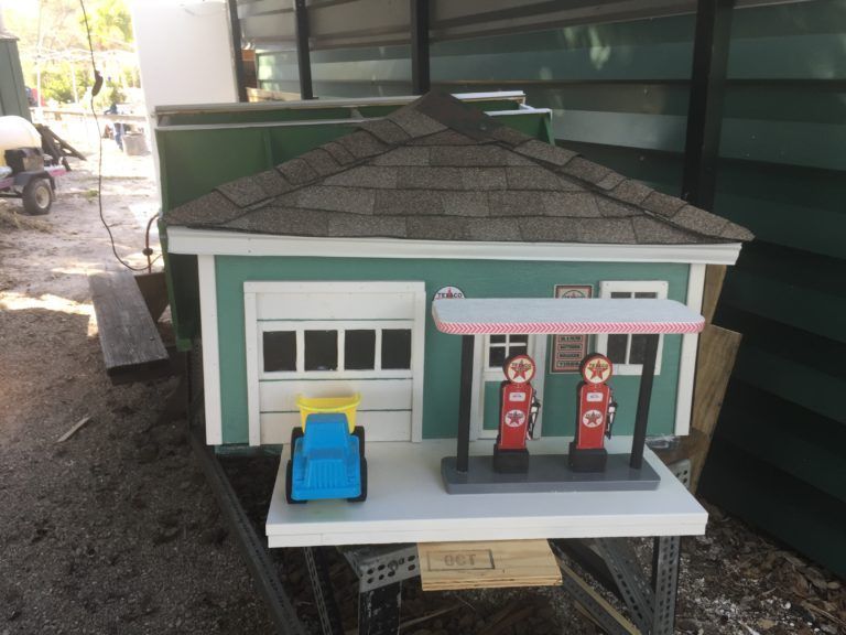 Toy gas station with red and white pumps, blue and yellow car, and green building with gray roof.