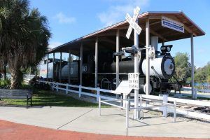 Steam locomotive under a canopy, on display next to a white picket fence and bench in a park.