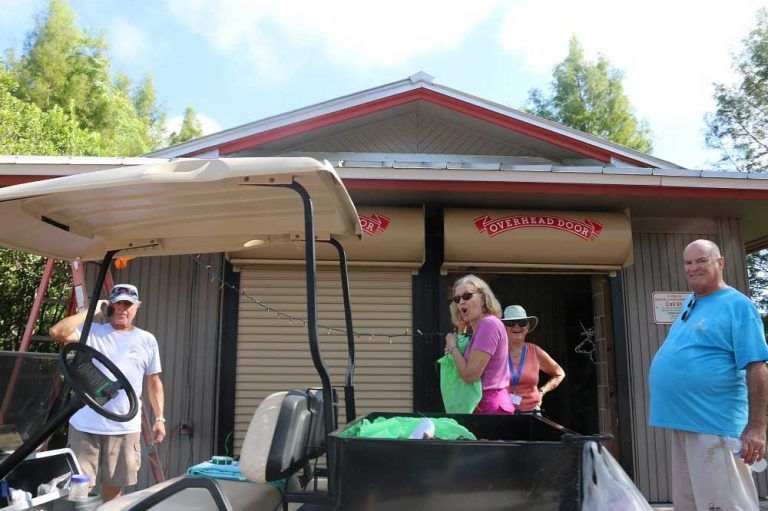 Four people stand near a golf cart in front of a small building. The building has a roll-up door with red lettering.