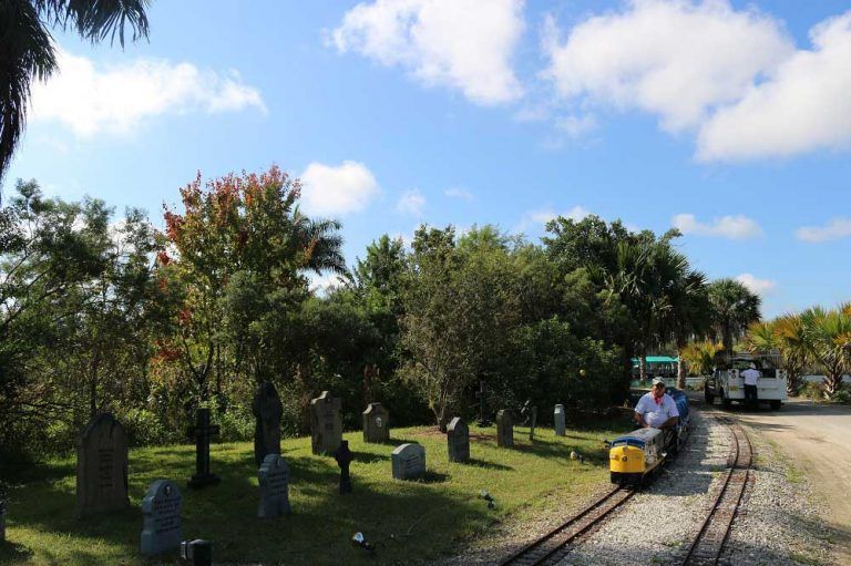 A miniature train travels past a graveyard. The train is yellow, with a man driving it. Overcast, sunny day.