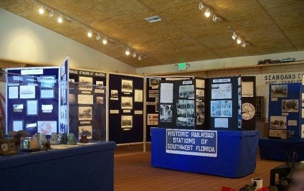 An exhibit hall displays historical photos and documents about historic railroad stations in Southwest Florida. The display boards sit atop blue tables, and the room has track lighting.