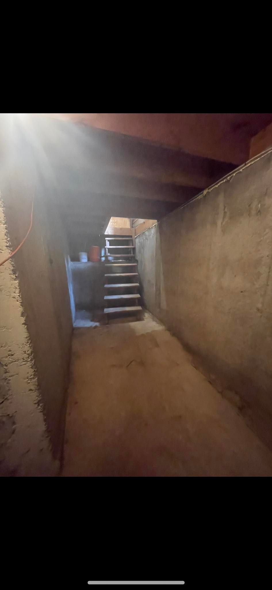 A narrow, dimly lit basement corridor with concrete walls, exposed ceiling joists, and a set of wooden stairs.