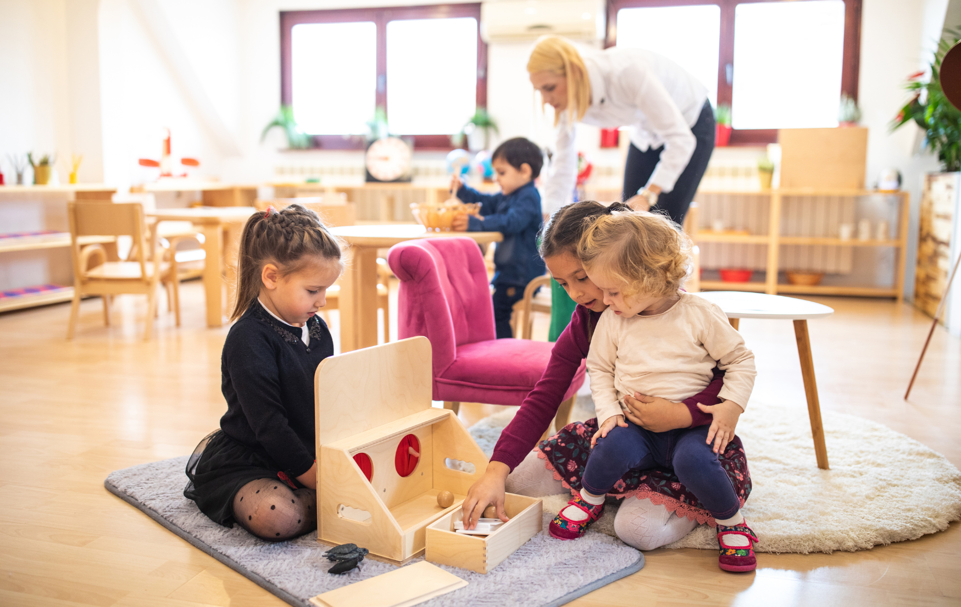 A teacher supervises children playing with wooden learning toys on a rug in a bright, organized classroom.