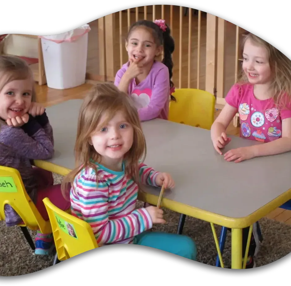 Four children sit around a gray table in a classroom, smiling and interacting during a group activity.