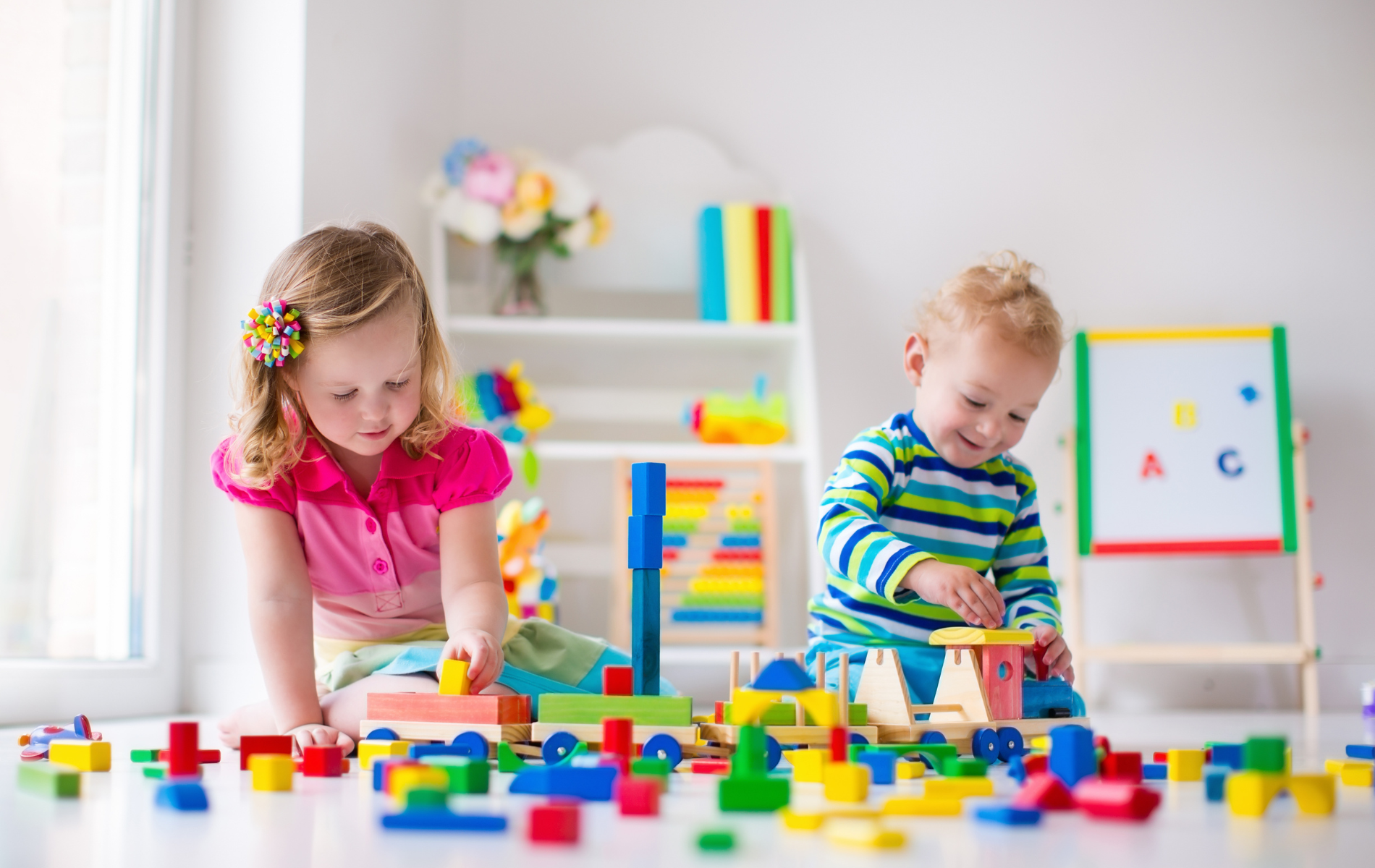 Two children sitting on the floor playing with colorful wooden building blocks in a bright room with toys.
