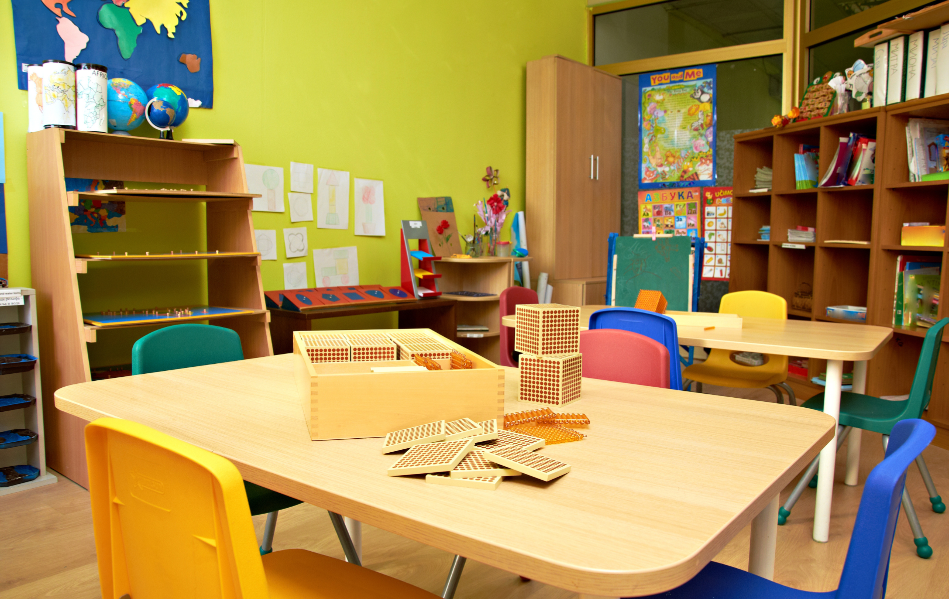 A bright, colorful preschool classroom featuring wooden tables, small chairs, toy storage, and educational wall posters.