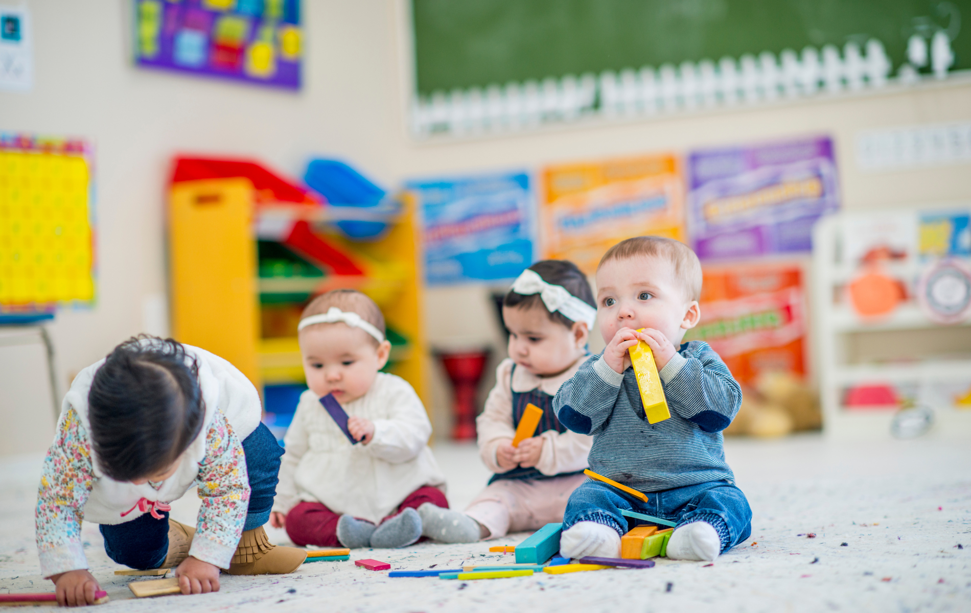 Three toddlers sitting on a playmat in a classroom, playing with colorful building blocks.