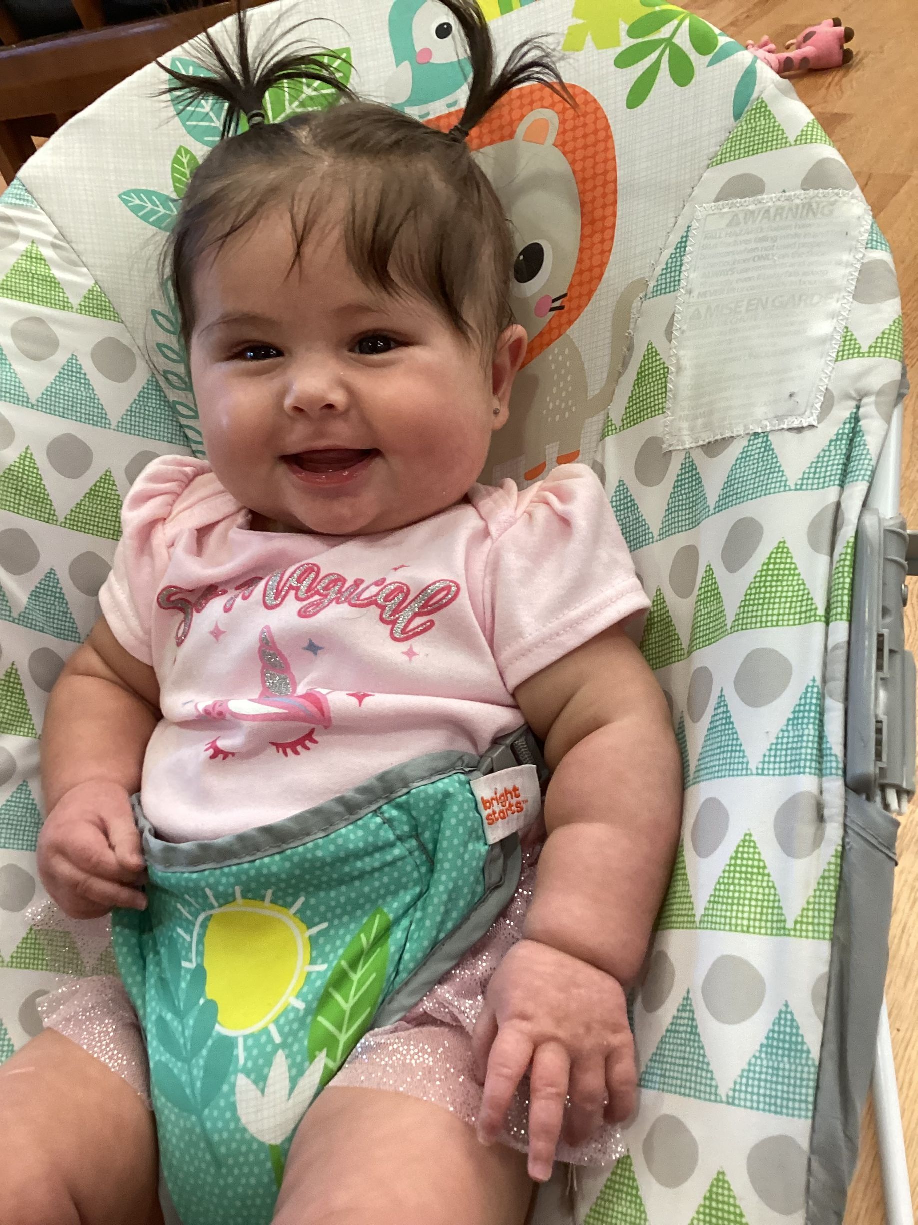 A smiling baby with two small ponytails sitting in a patterned infant bouncer, wearing a pink outfit and a green cover.