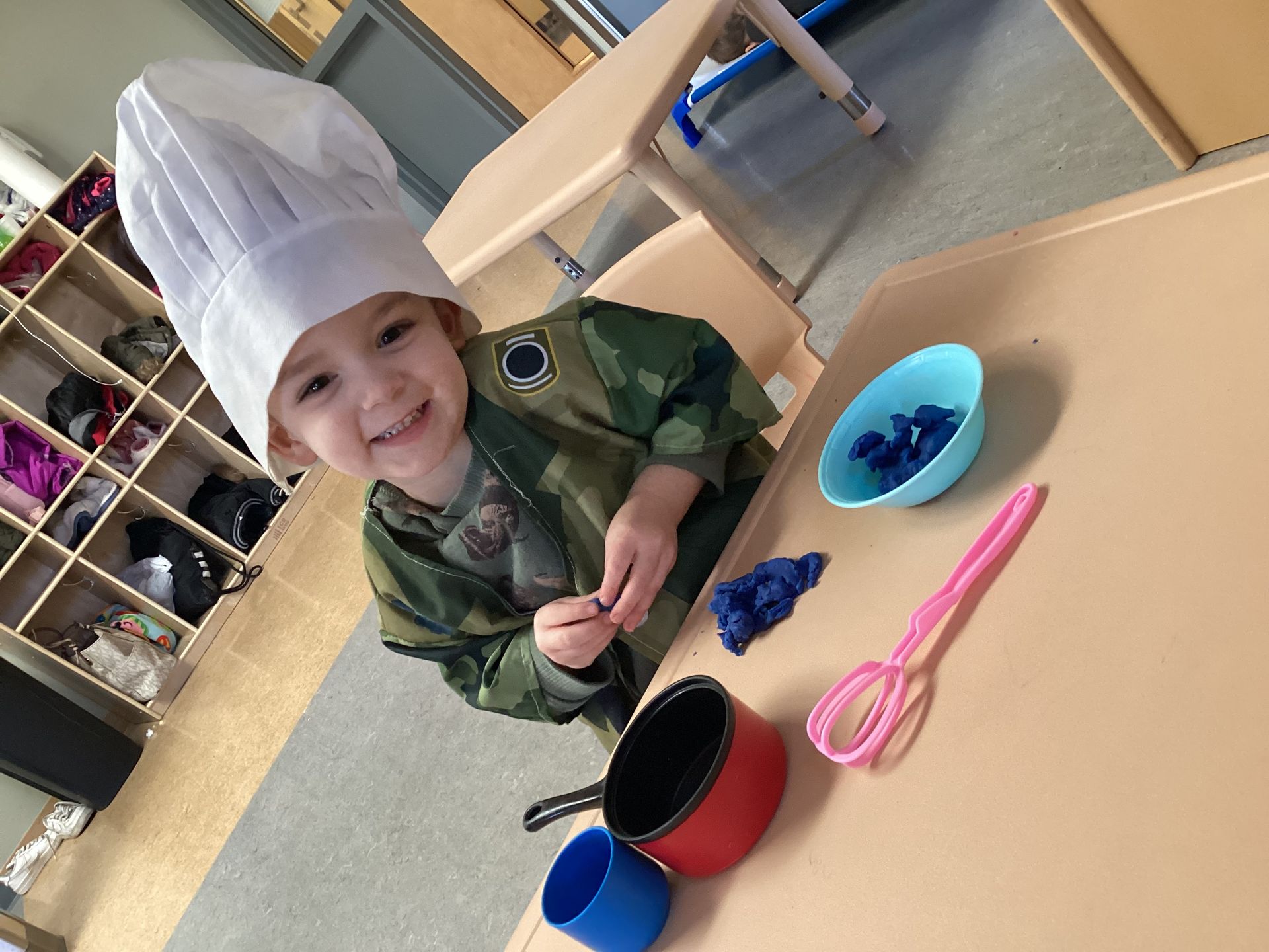 A toddler wearing a chef’s hat smiles while playing with a toy pot, blue bowl, whisk, and playdough at a table.