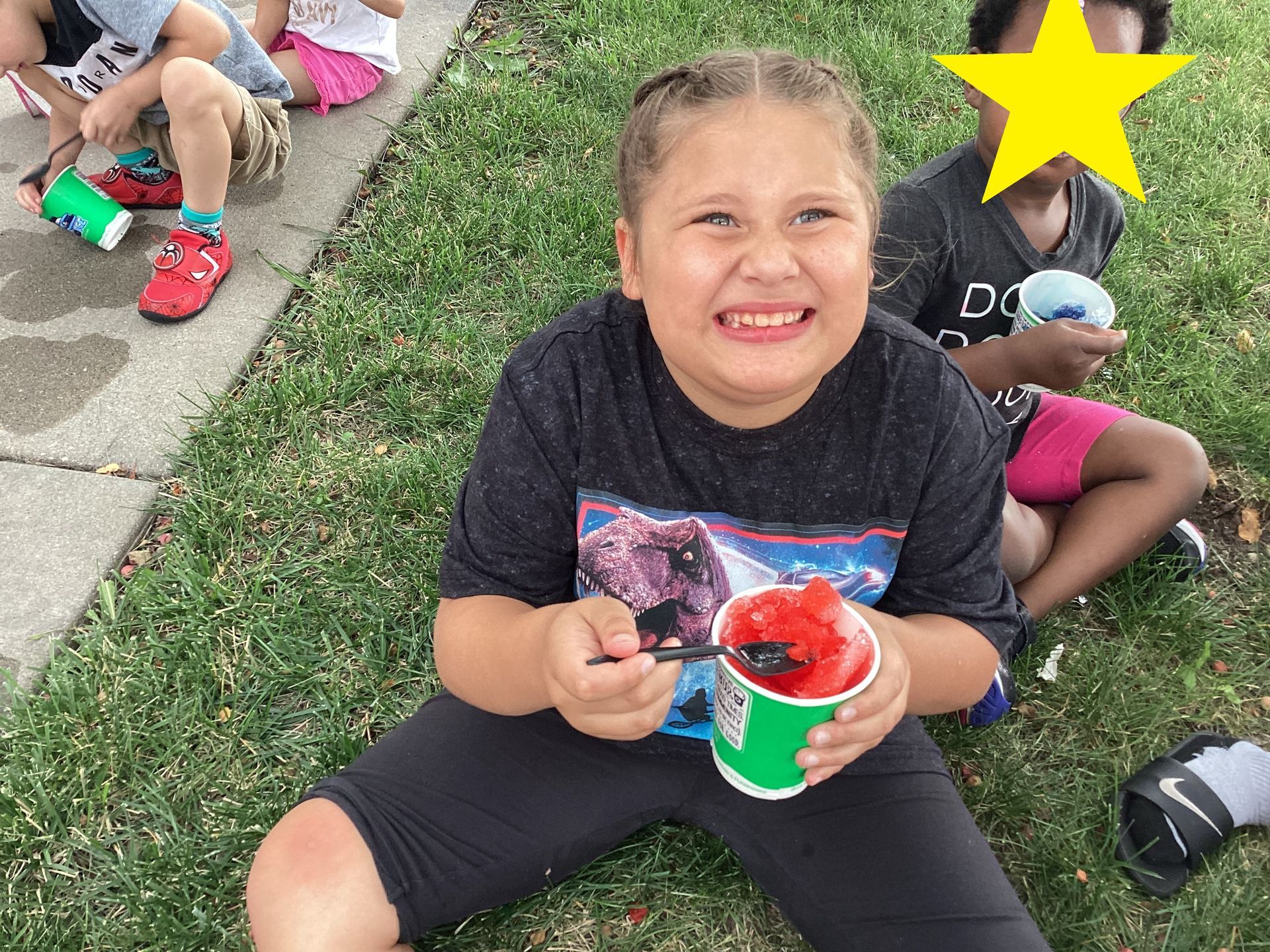 A person smiling while sitting on the grass outdoors and eating a red shaved ice treat from a green cup.