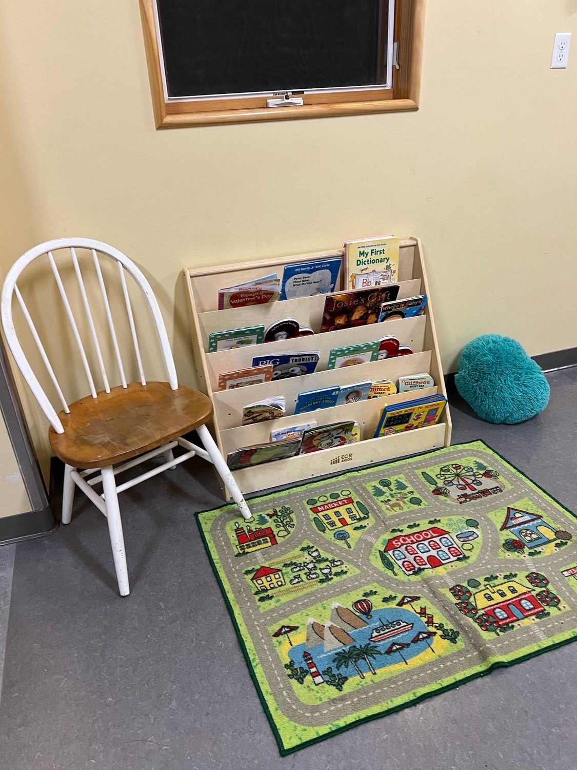 A small wooden chair next to a toy-filled bookshelf and a road-themed play rug on a floor near a window.