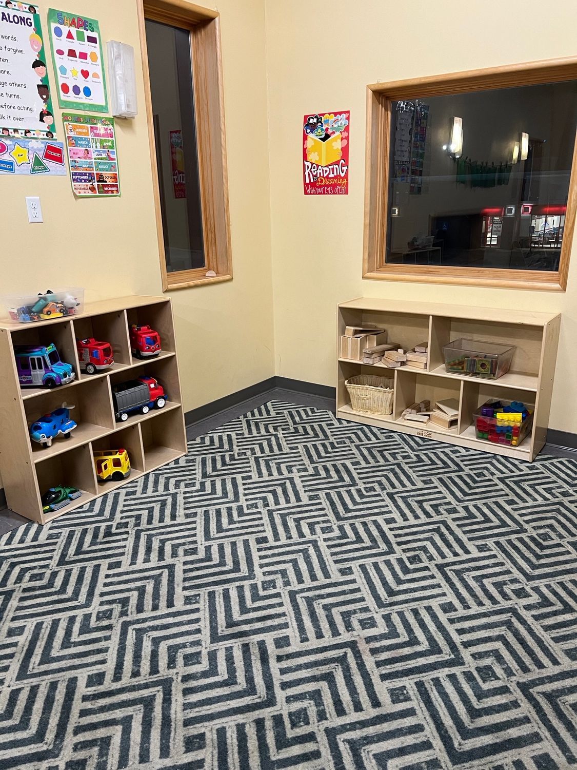 A colorful classroom corner features two wooden cubby shelves with toys, a patterned rug, and wall posters.