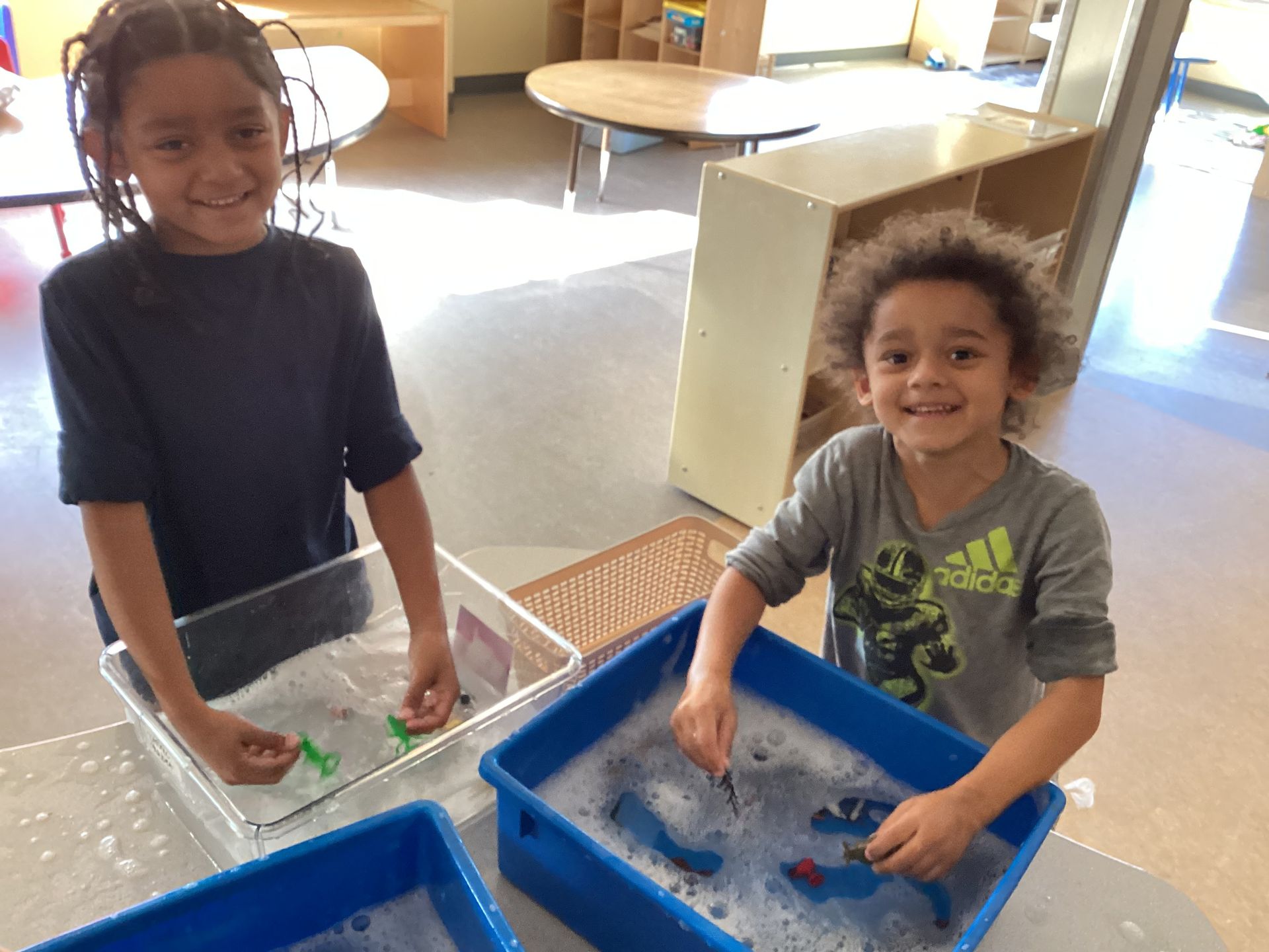 Two children smile while washing small plastic toy animals in soapy water-filled bins at a preschool table.