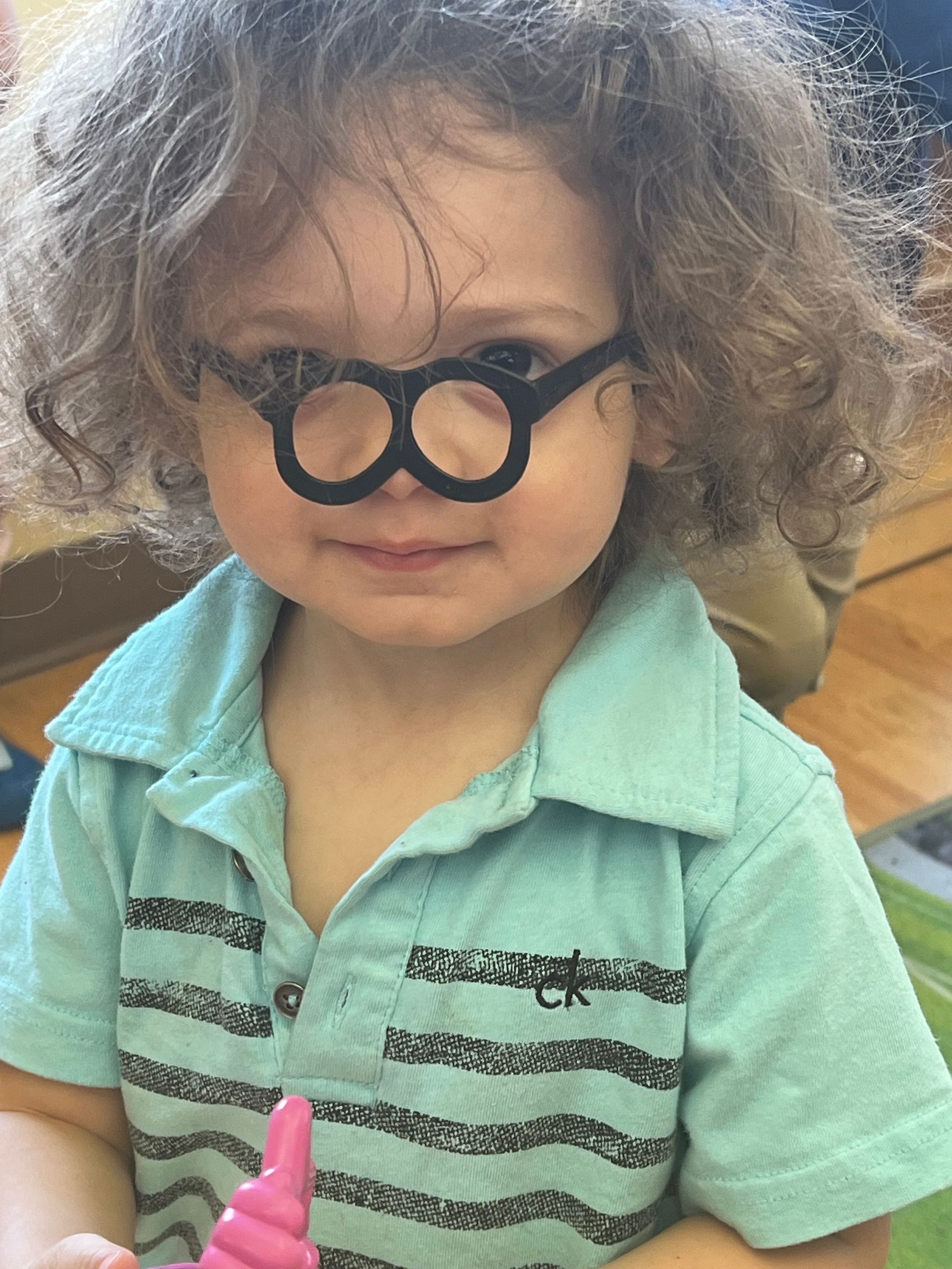 A toddler with curly hair wears black paper glasses over their nose, wearing a light blue polo shirt with stripes.