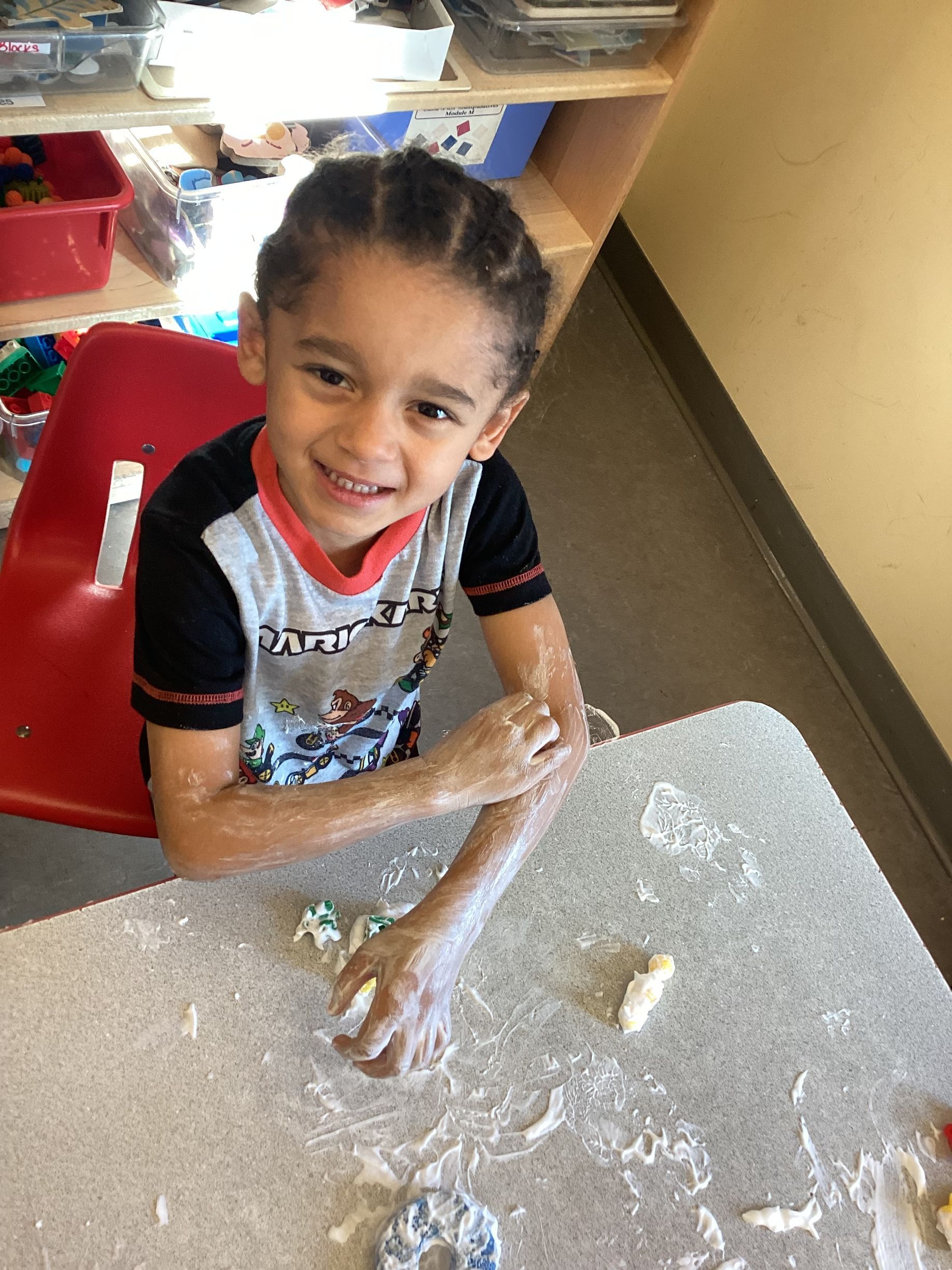 A smiling child in a gray and black shirt sits at a desk covered in white modeling clay, with clay on their hands and arms.