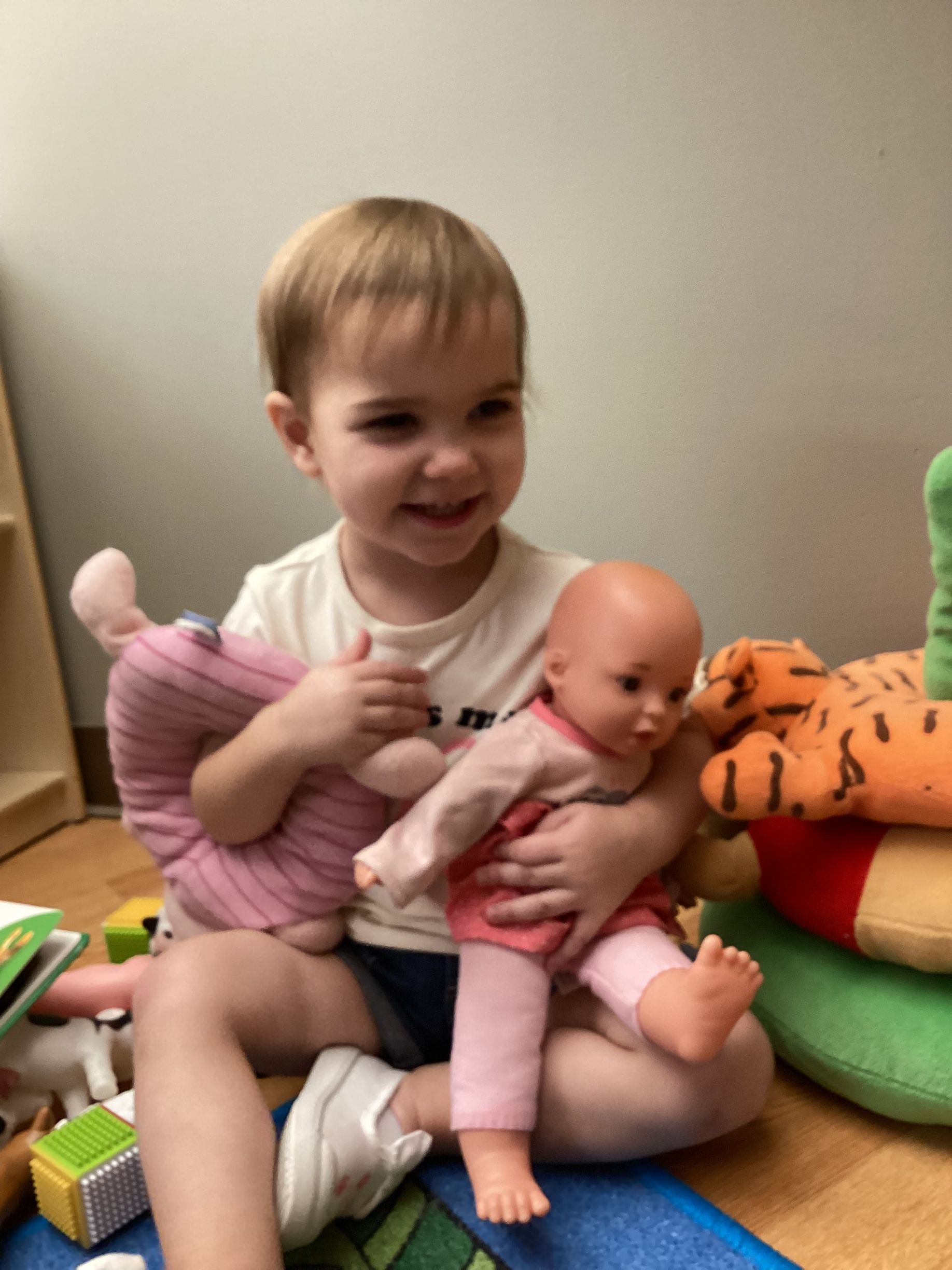 A child sits on the floor, smiling while holding a doll and a pink stuffed animal, surrounded by various toys.