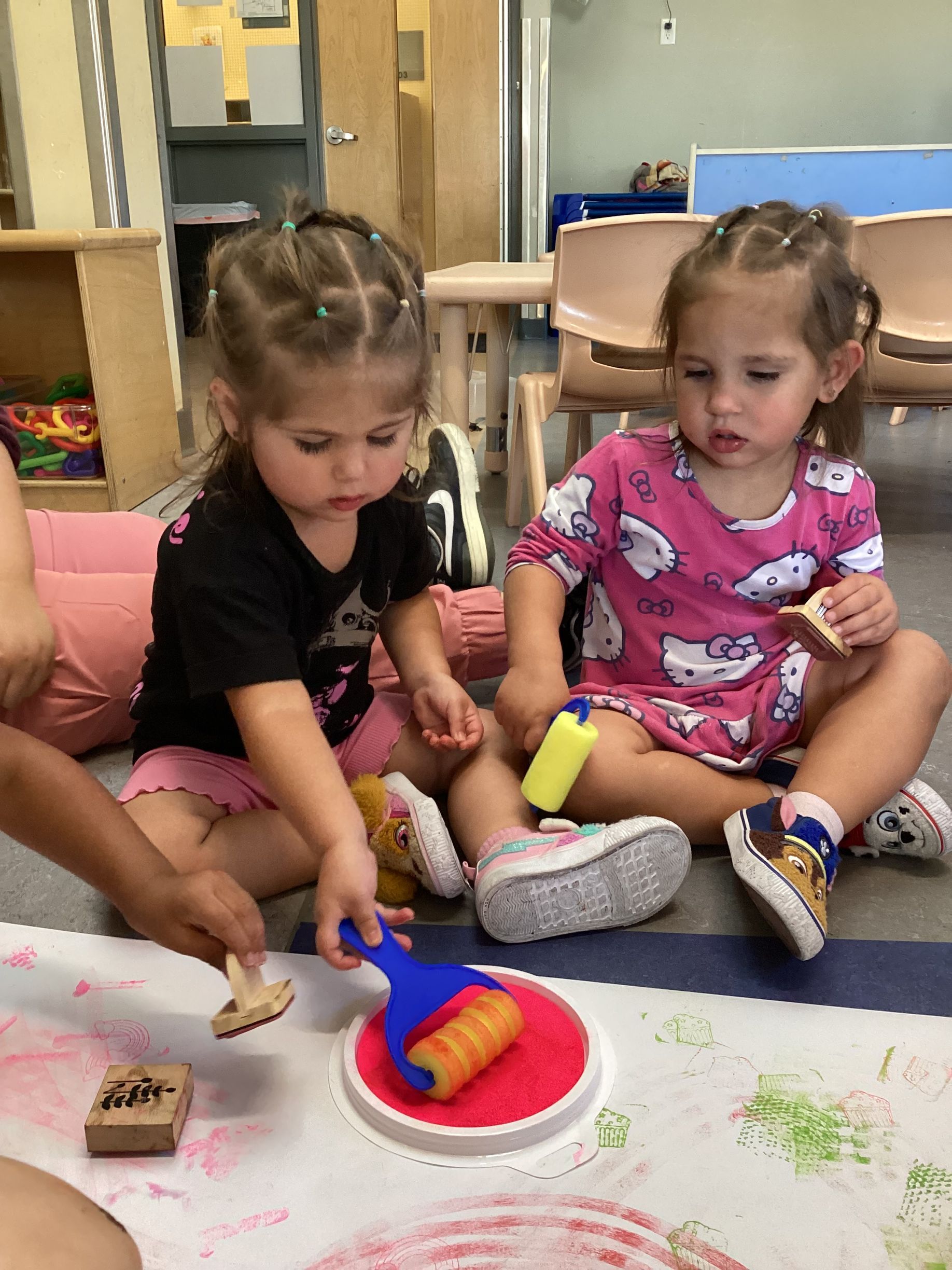 Two young children sitting on a floor, using paint rollers and stamps on paper in a classroom setting.