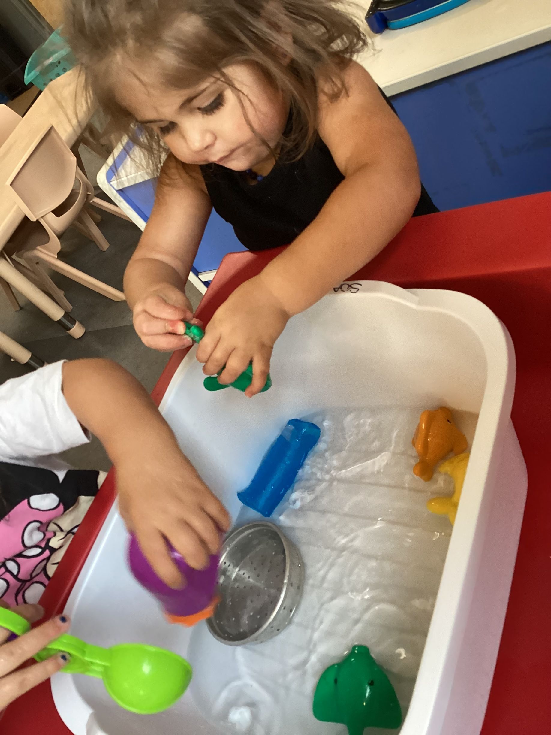 Two children play with colorful plastic scoops, a strainer, and small toys in a white water table.