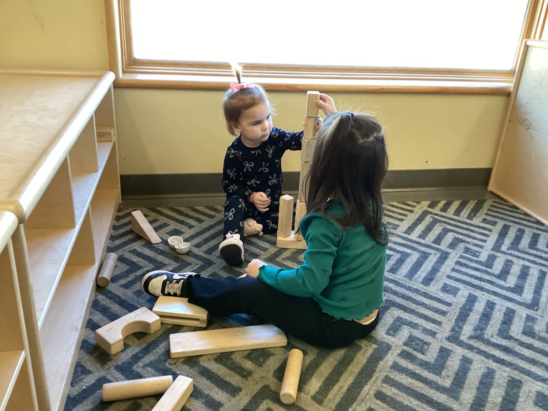 Two children sit on a patterned carpet near a wooden shelf, focused on stacking wooden blocks together.