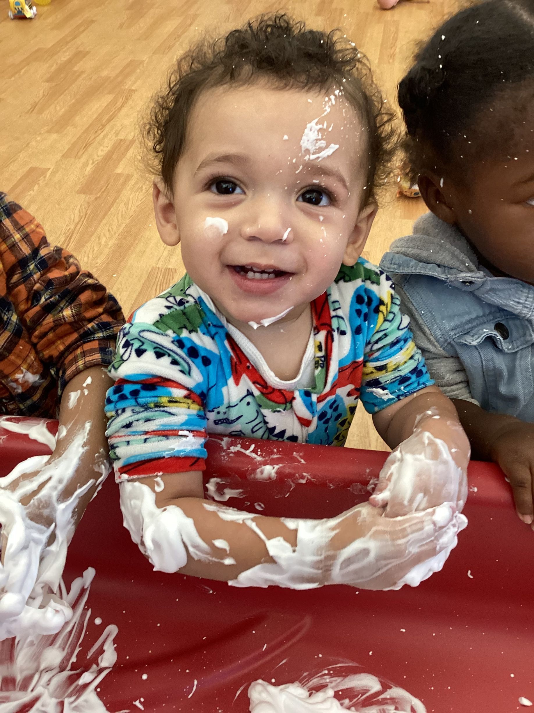 A toddler with hands covered in white foam smiles while playing at a red sensory table.
