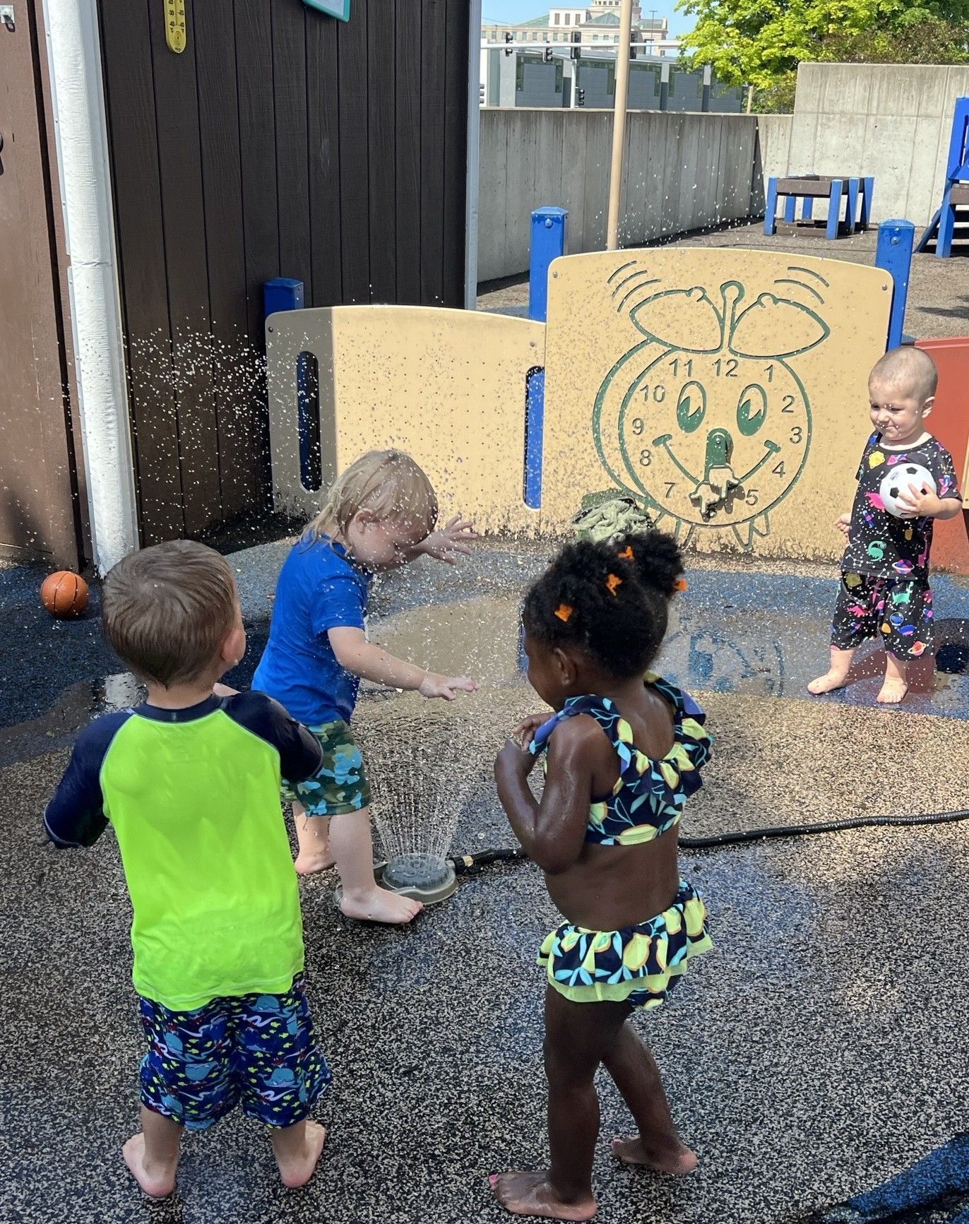Children in swimwear playing in a water spray area with a clock-themed wall panel at a playground.