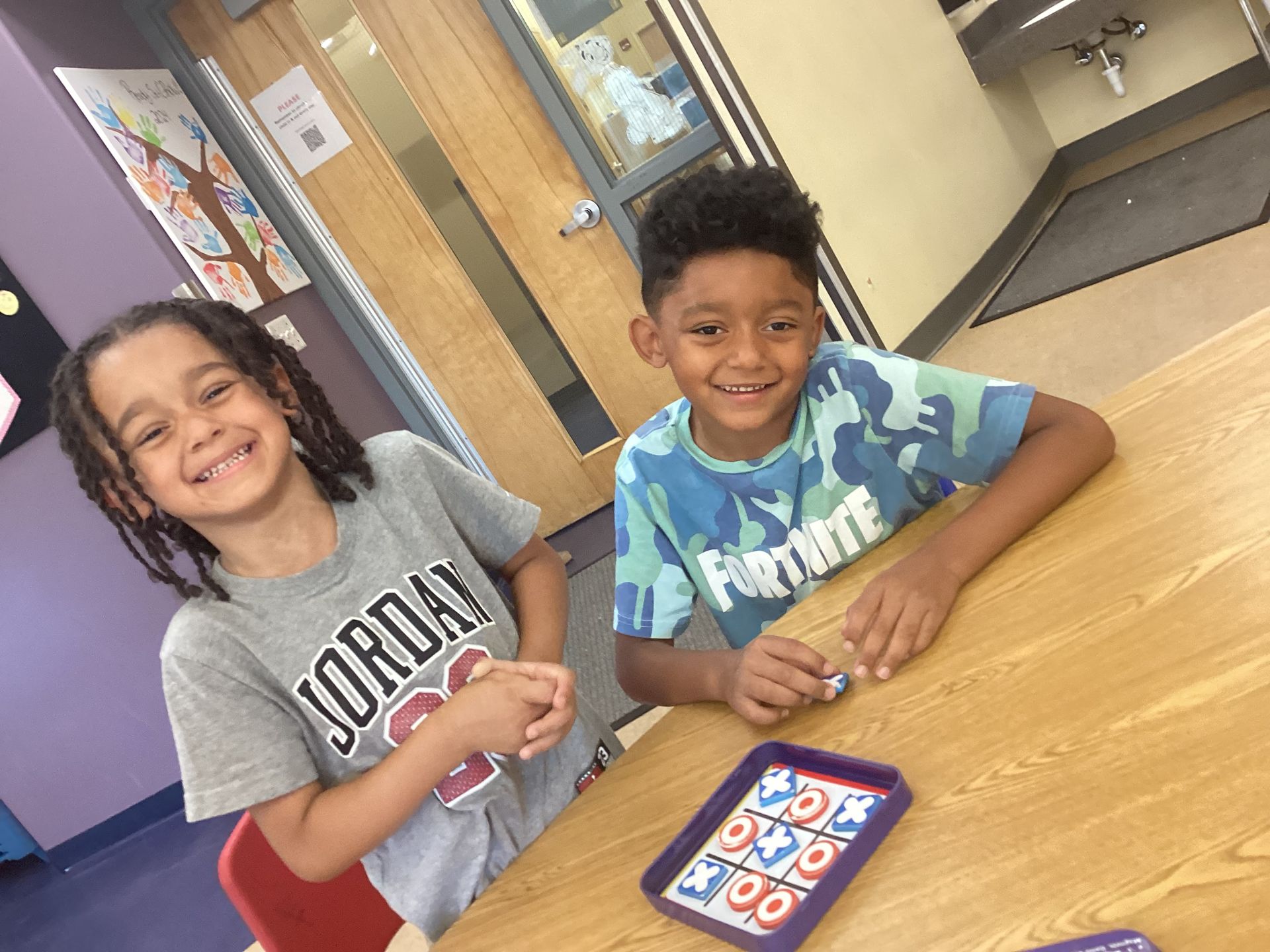 Two children smiling while playing a Tic-Tac-Toe game at a table in a classroom.