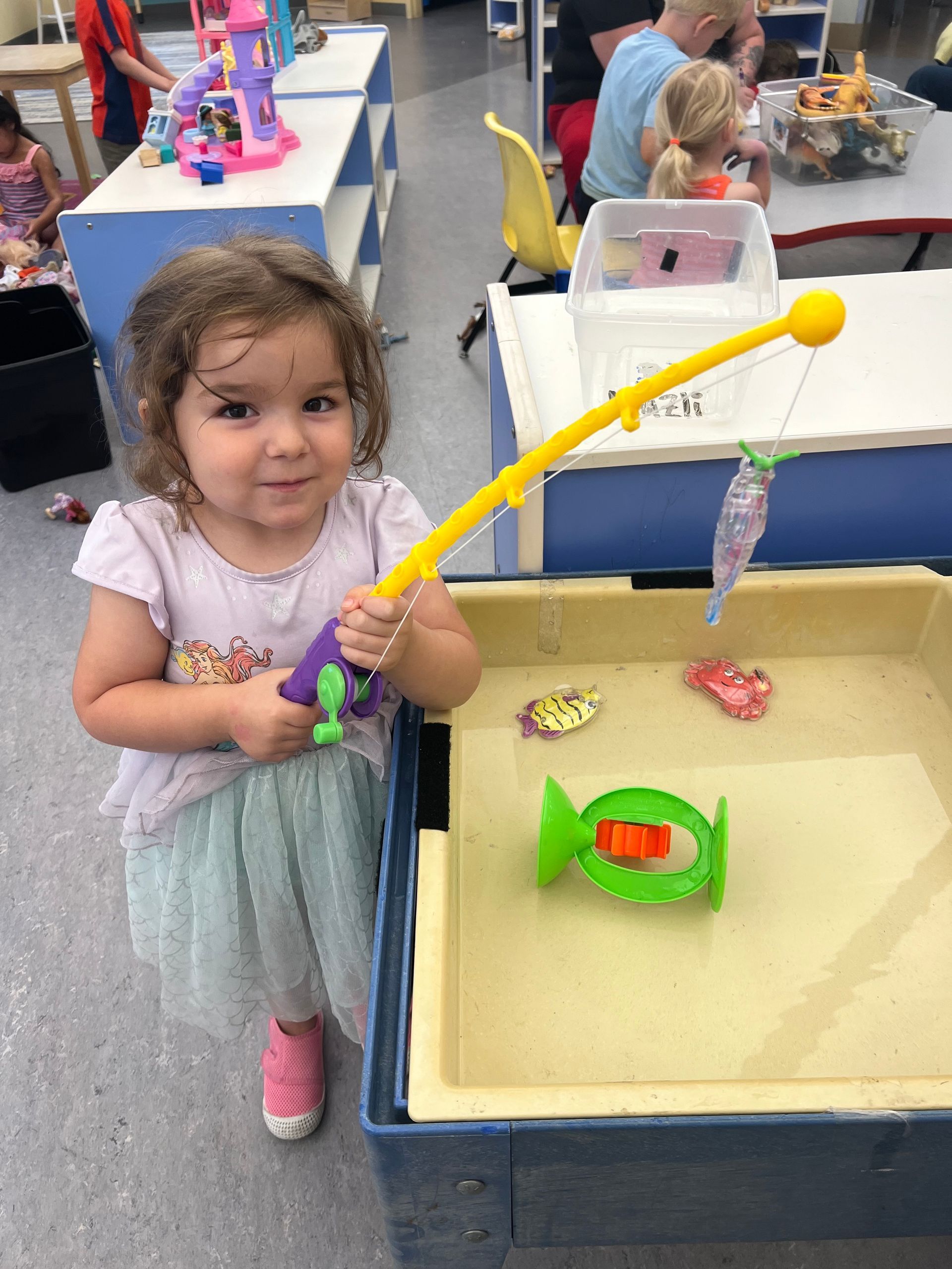 A young child smiles while holding a toy fishing rod above a water table with floating plastic sea creatures in a room.