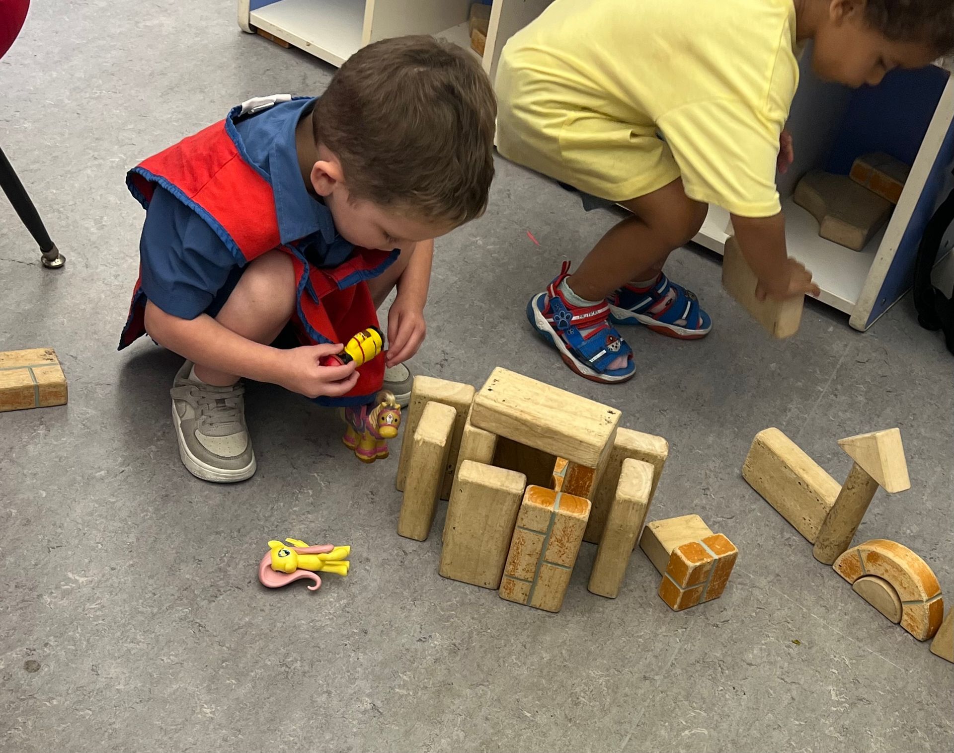 Two children play with wooden building blocks and small figurines on a grey floor in a classroom setting.