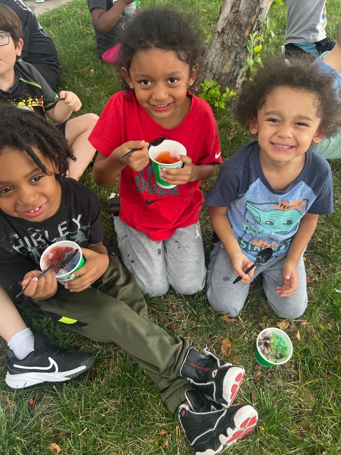 Three children sit on grass outdoors, smiling while holding and eating from small paper cups of frozen treats.