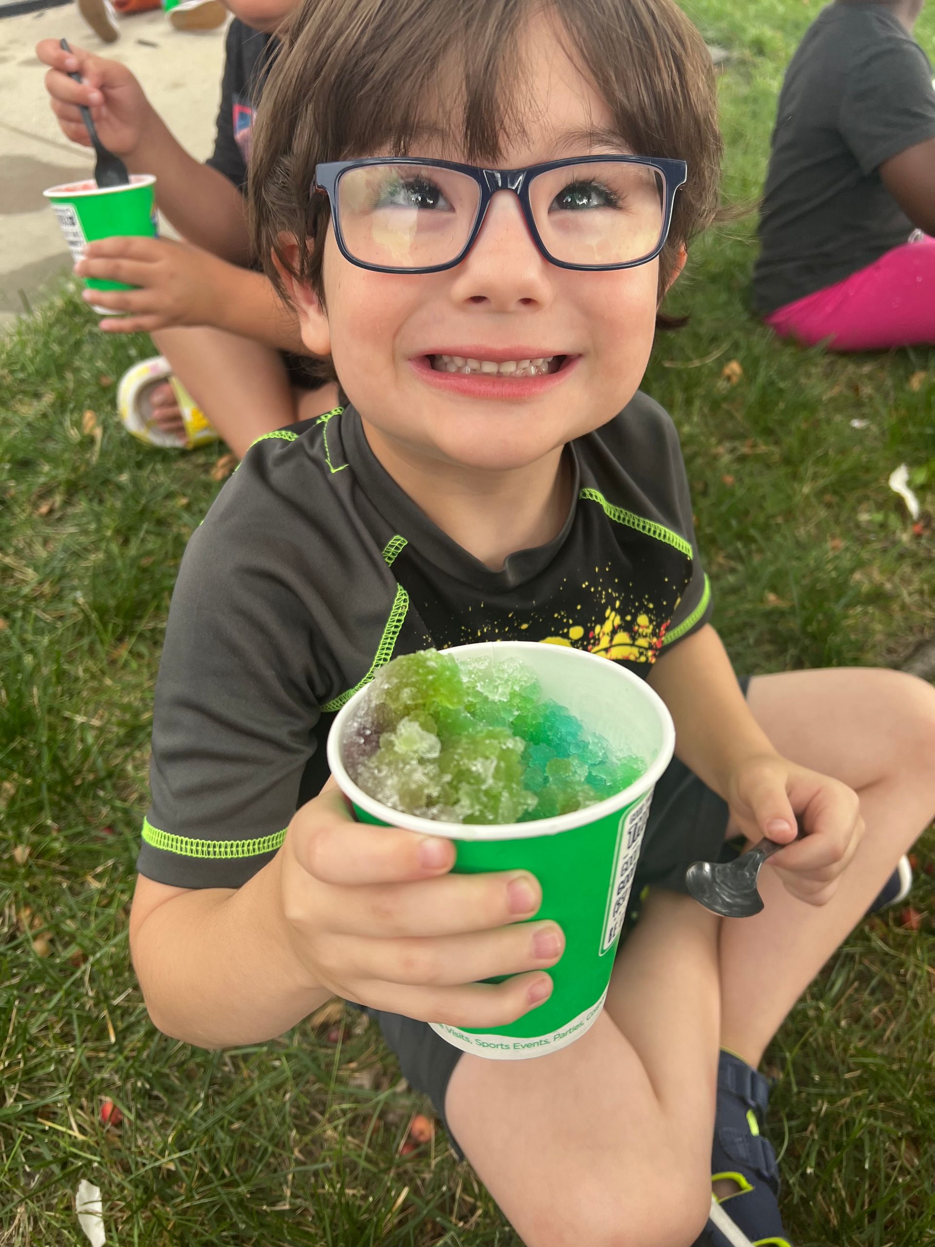 A child with glasses smiles while holding a cup of colorful shaved ice while sitting on the grass.