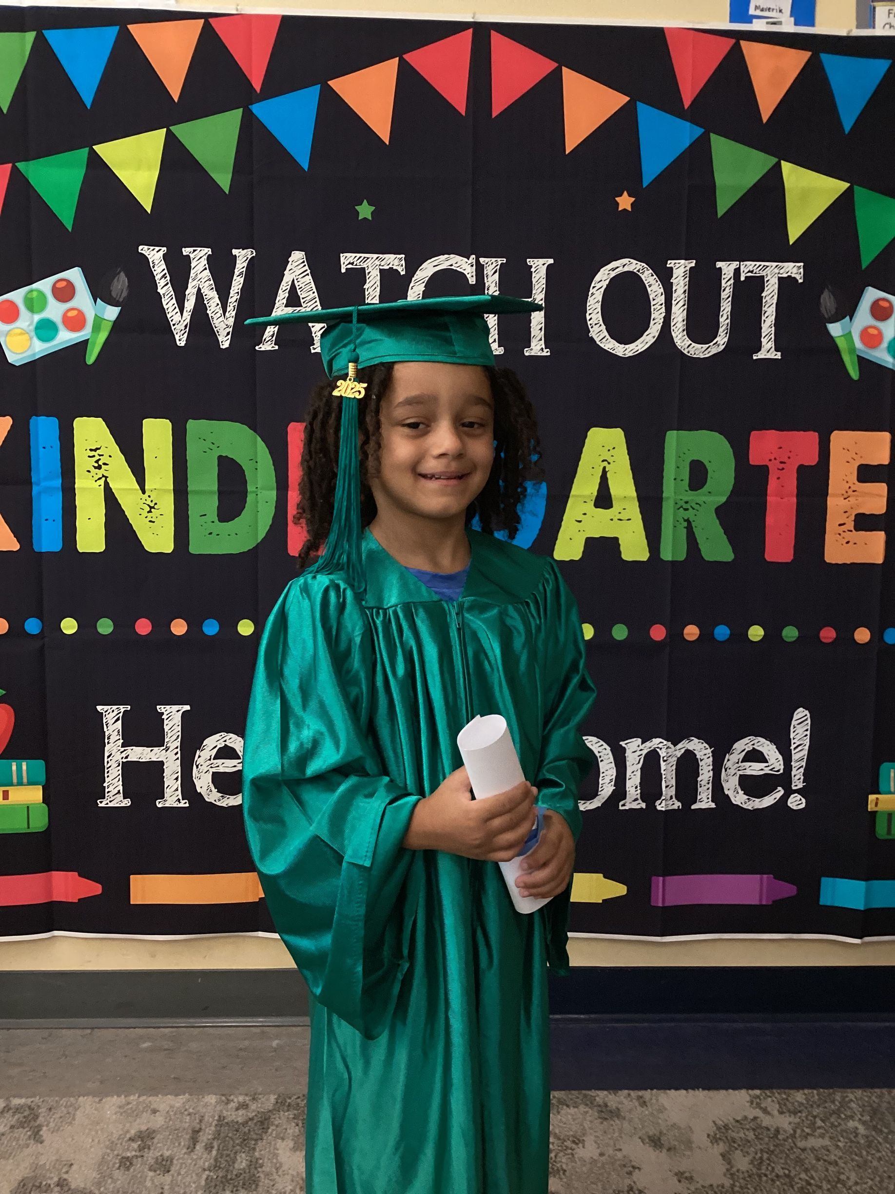 A student in a green graduation cap and gown holding a diploma in front of a “Watch Out Kindergarten” banner.