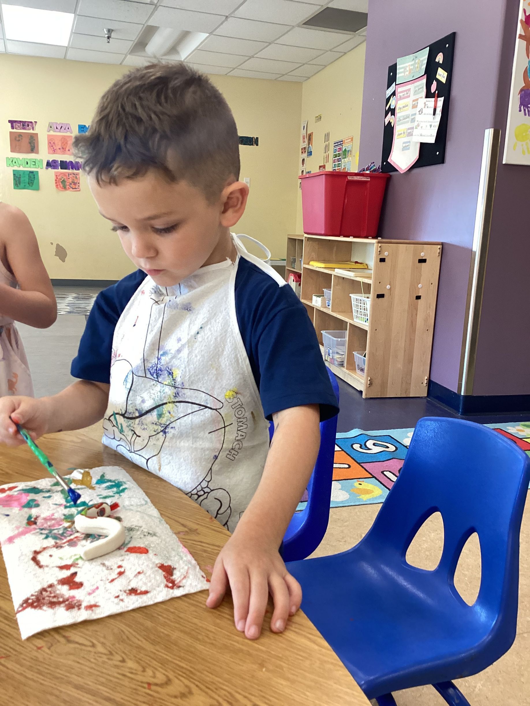 A person wearing a paint-splattered apron sits at a table in a classroom, painting a small, white clay object.