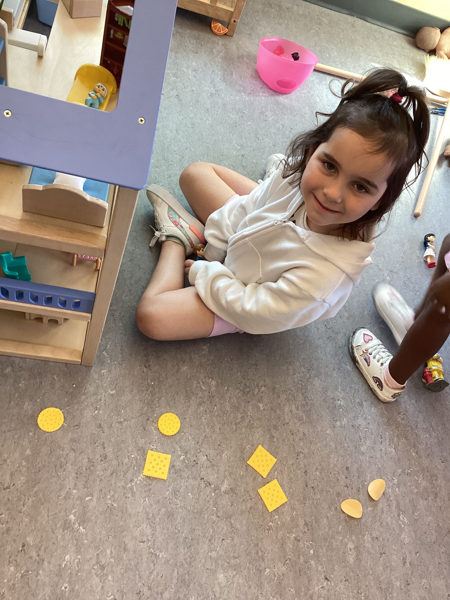 A child sits on the floor smiling, next to a dollhouse with yellow shapes arranged in a line on the carpet.