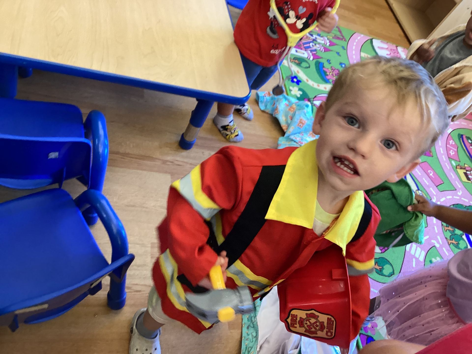 A child wearing a red and yellow firefighter costume holds a plastic fire helmet in a classroom setting.