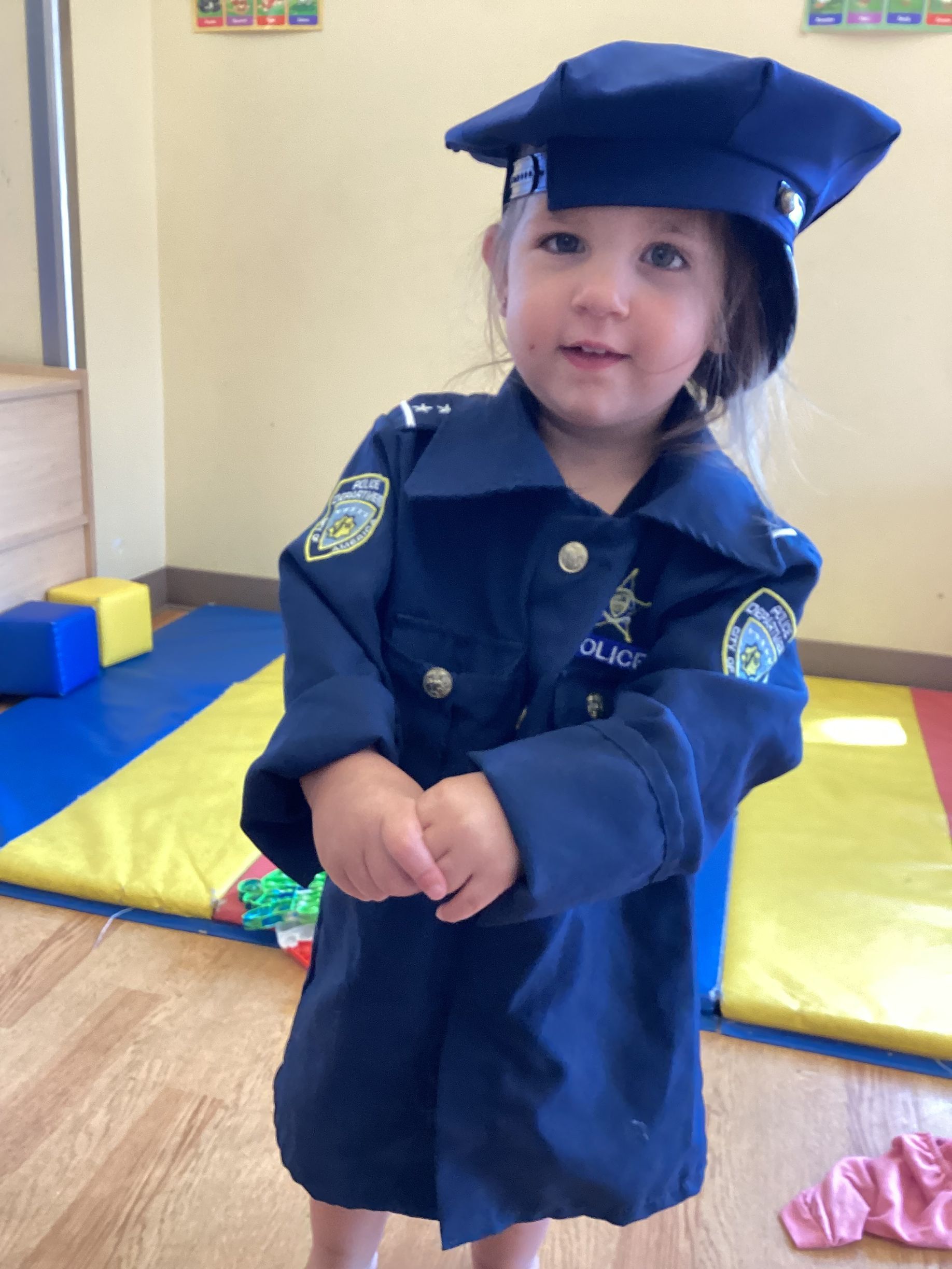 A young child in a blue police uniform and matching hat stands in a room with colorful floor mats.