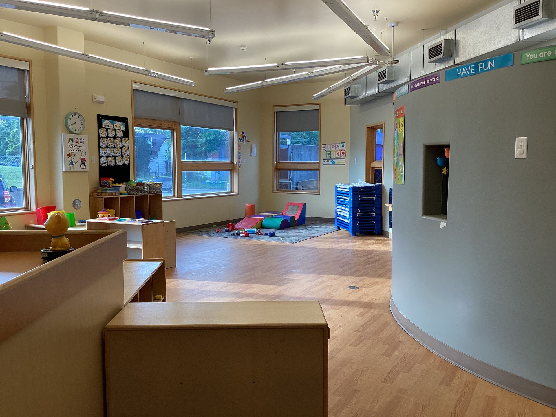 A brightly lit classroom with wooden floors, play areas, furniture, and windows looking out to the outdoors.