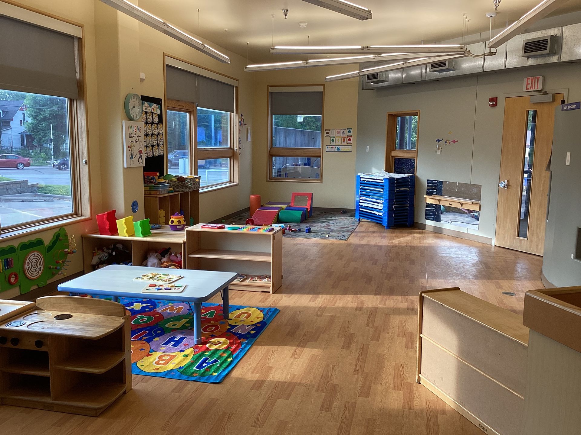 A brightly lit preschool classroom with wooden floors, a small blue table on a colorful rug, storage shelves, and windows.