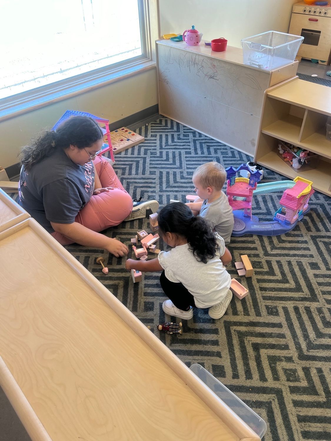 An adult and two children play with wooden toy furniture on a geometric patterned rug in a classroom.