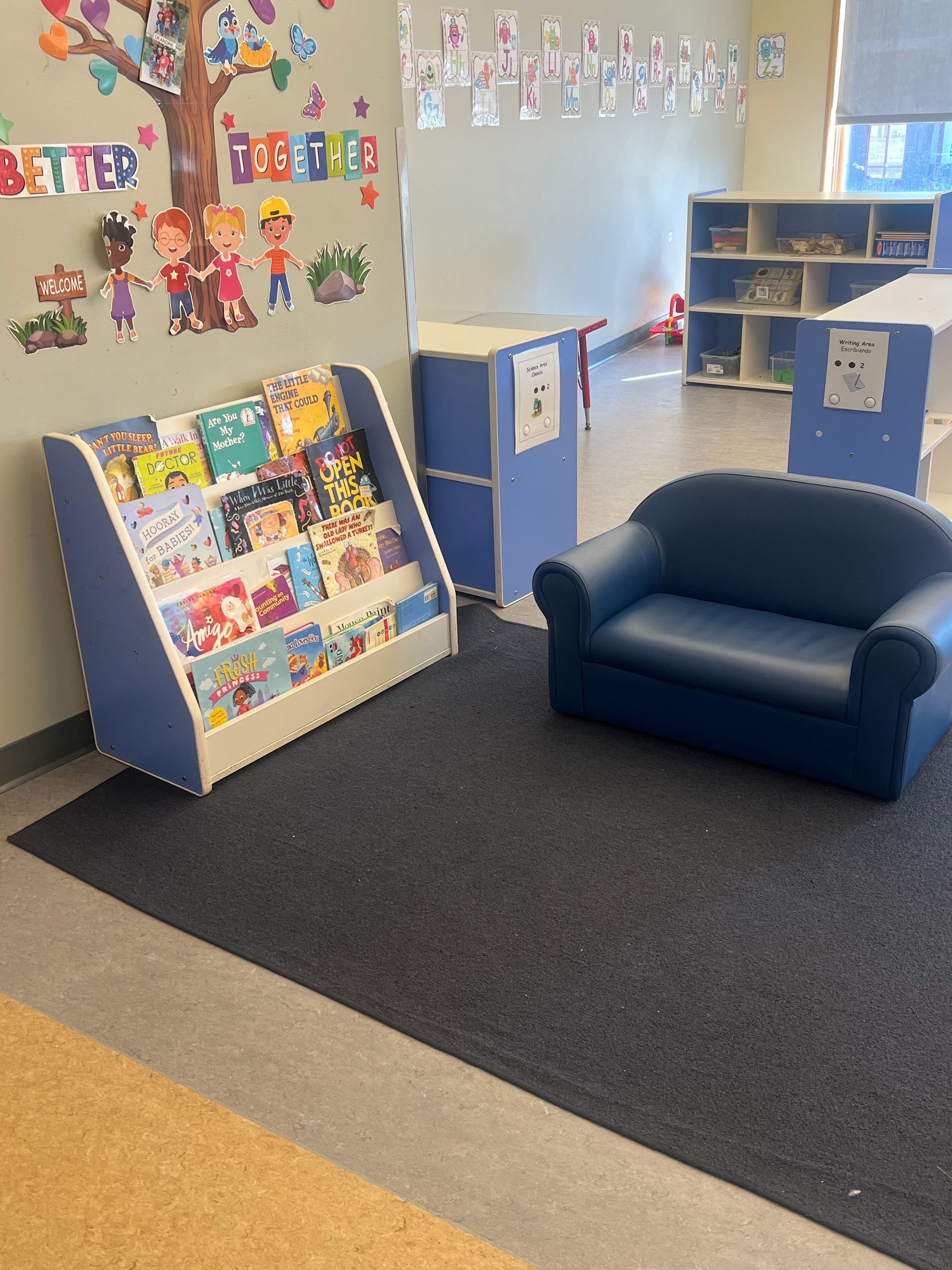 A cozy reading corner in a classroom with a blue armchair, a bookshelf filled with colorful books, and wall decorations.
