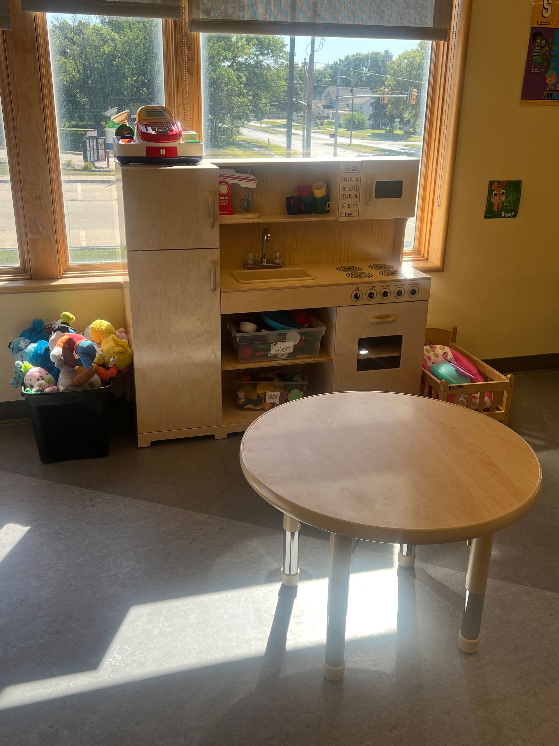 A play kitchen set with a refrigerator, a round light-wood table, and a bin of stuffed toys near a bright window.