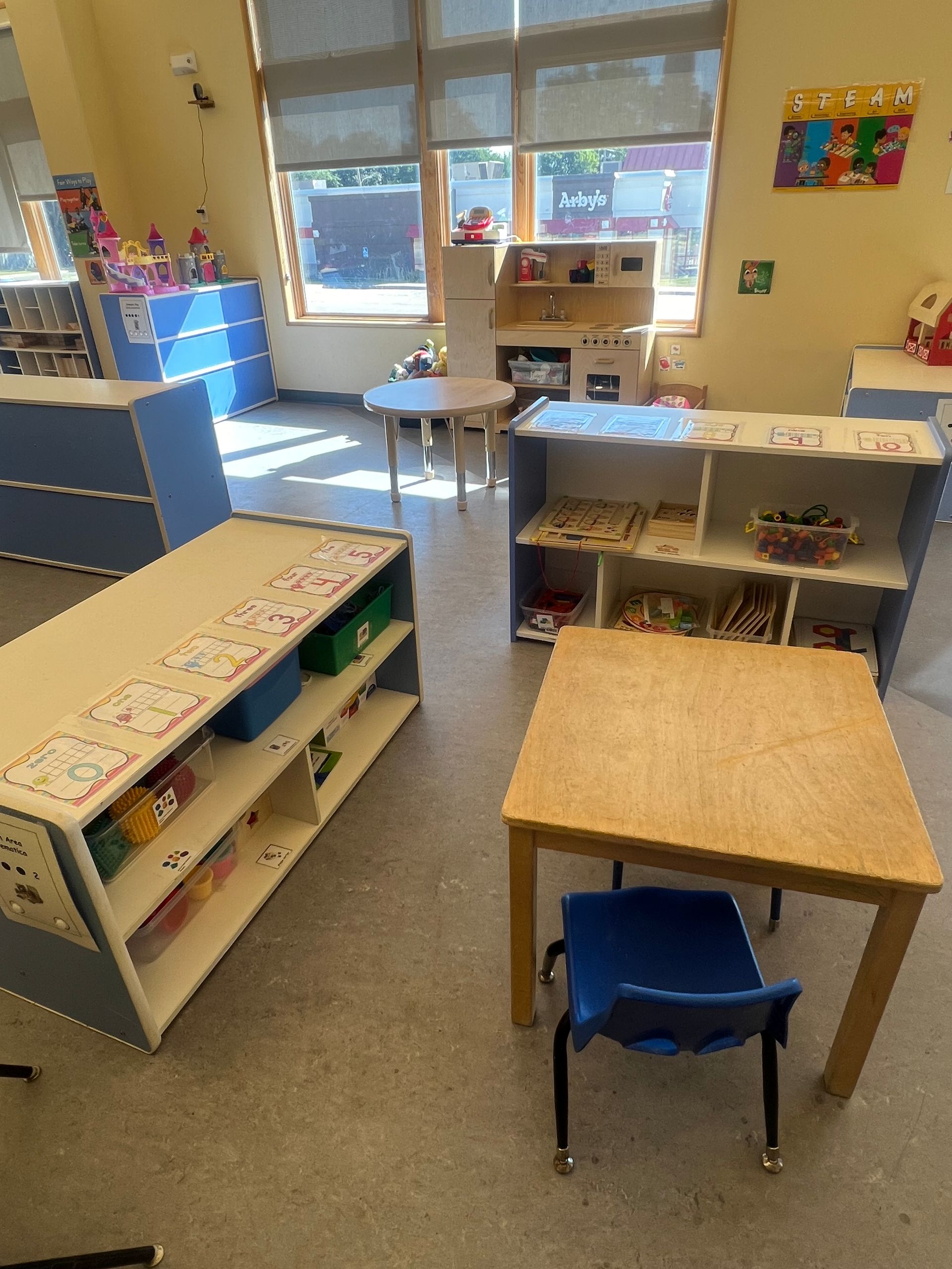 A sunny, organized preschool classroom with wooden tables, blue storage shelves, a play kitchen, and a small round table.