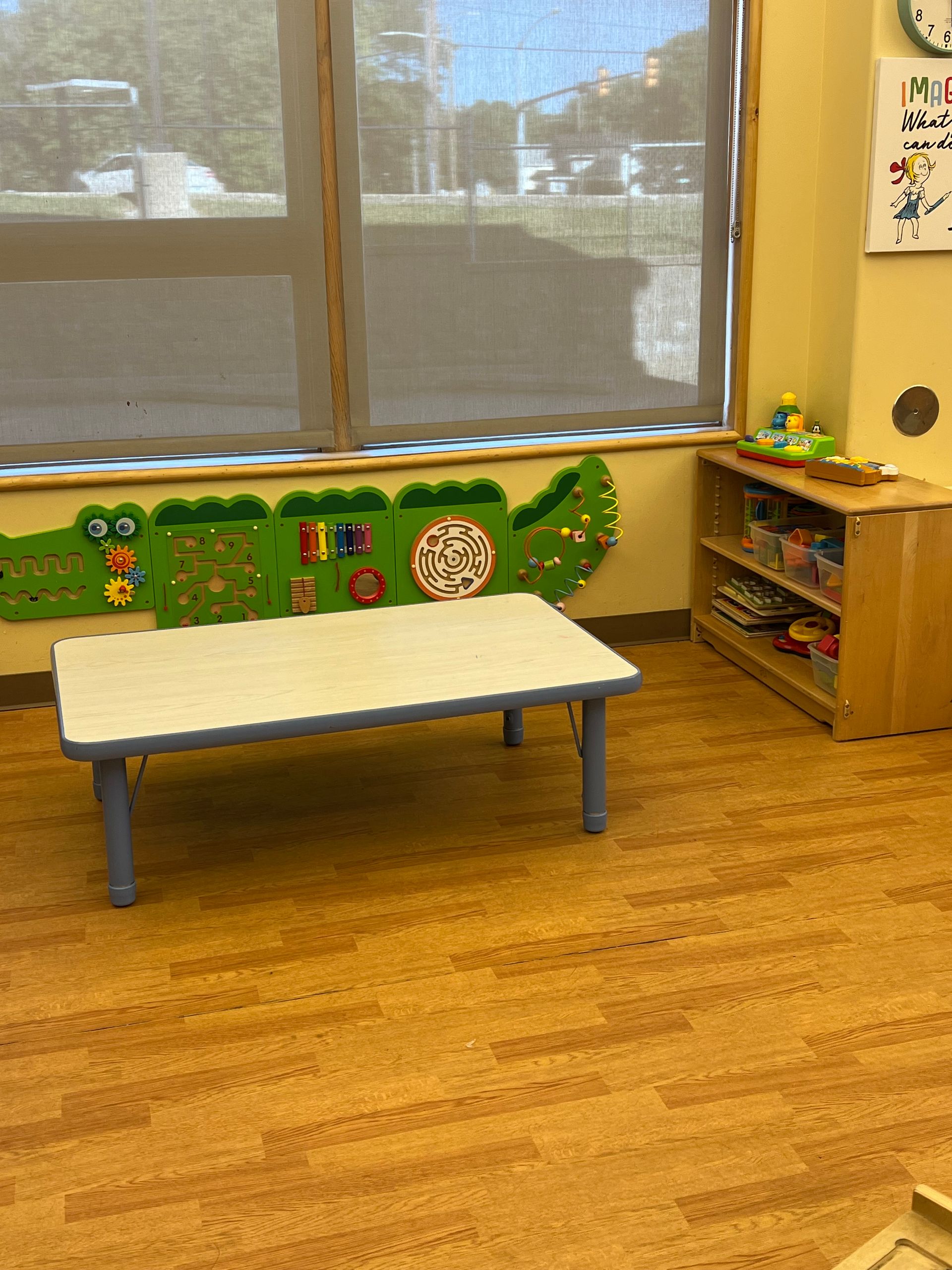 A classroom area with a light-colored table, a green crocodile-shaped wall activity board, and a small storage shelf.