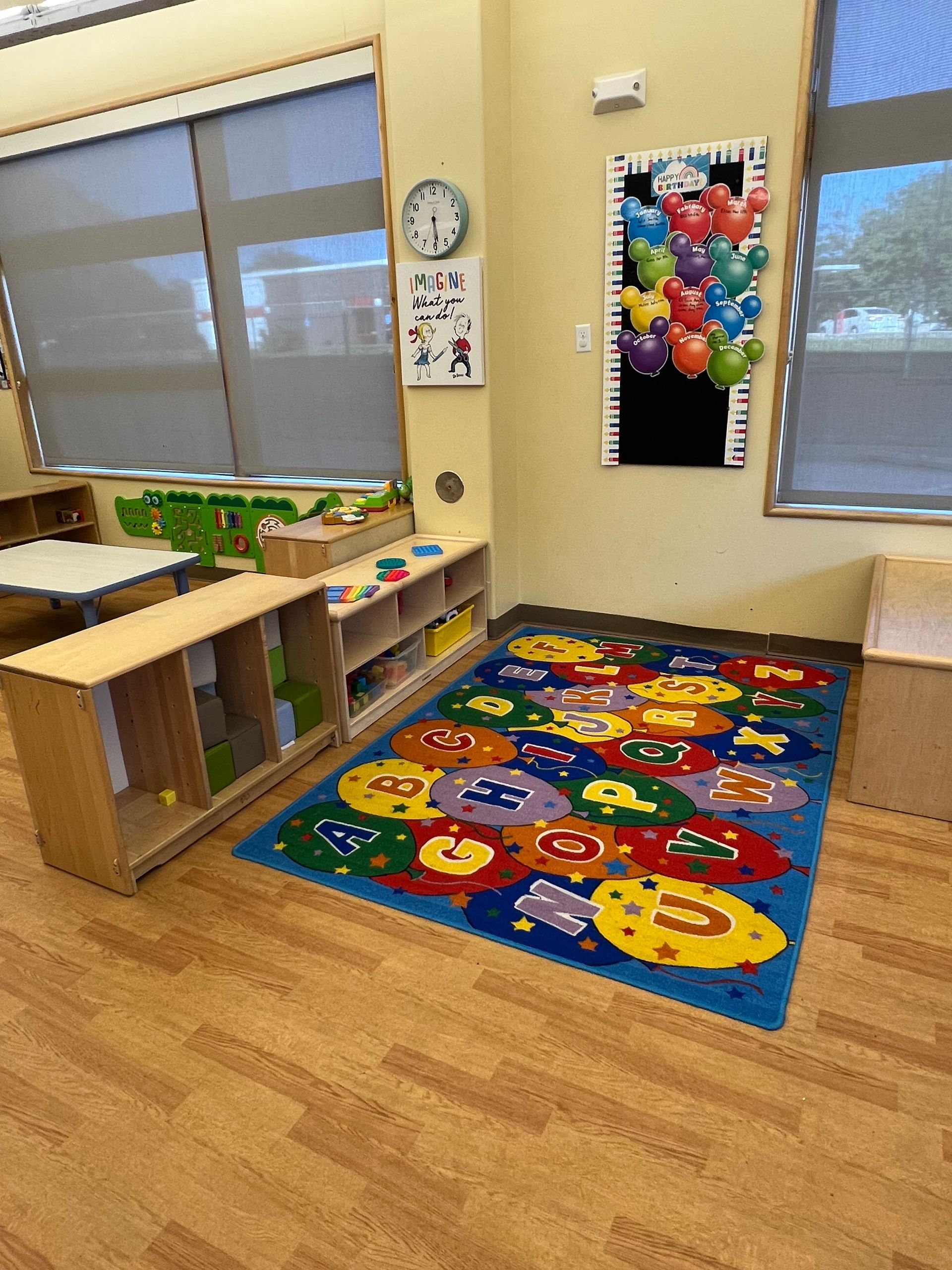 A classroom corner with a colorful alphabet rug, wooden storage shelves, and a Mickey Mouse-themed balloon wall display.