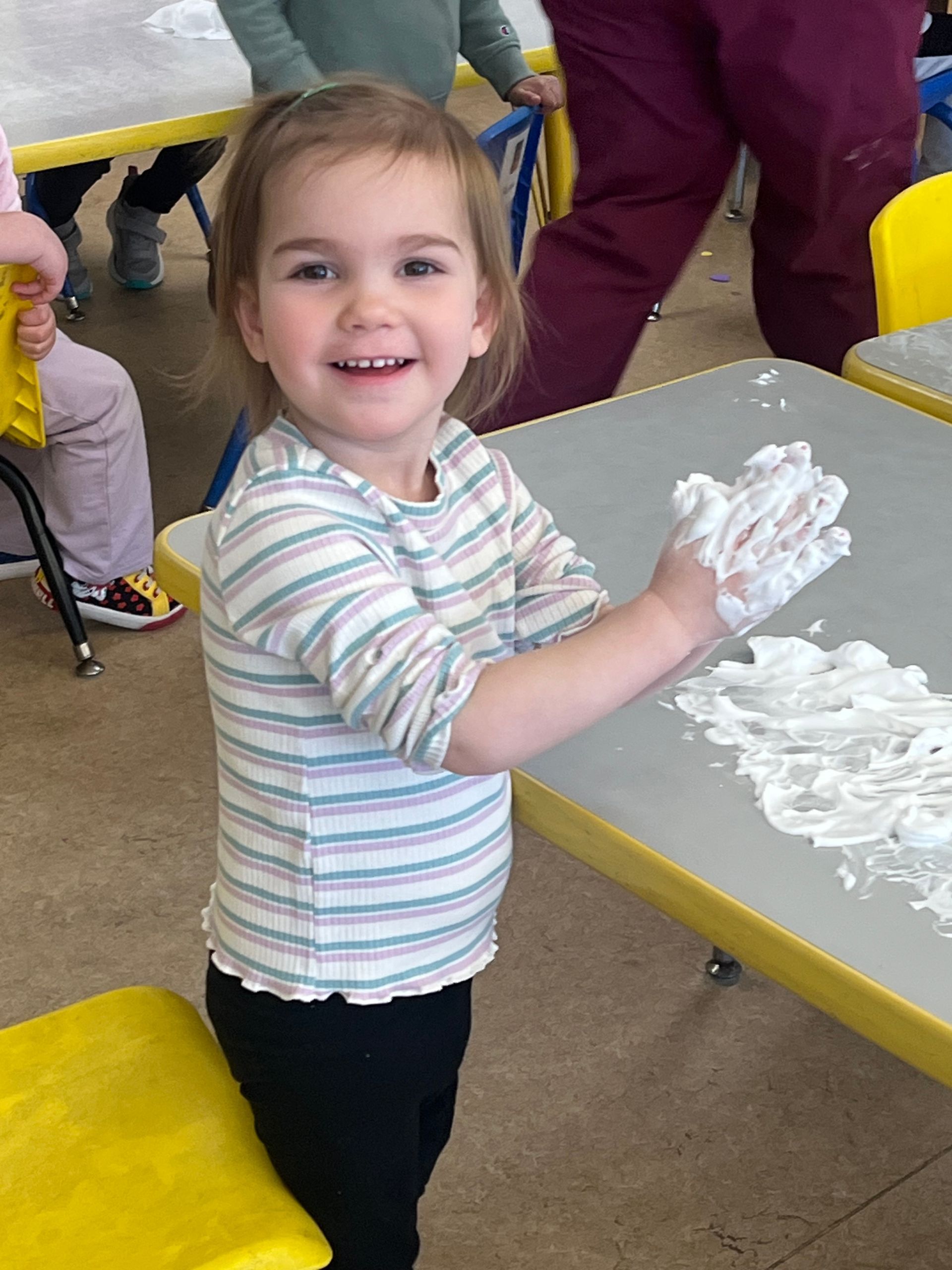 A smiling child wearing a striped shirt plays with white foam on a table in a classroom.