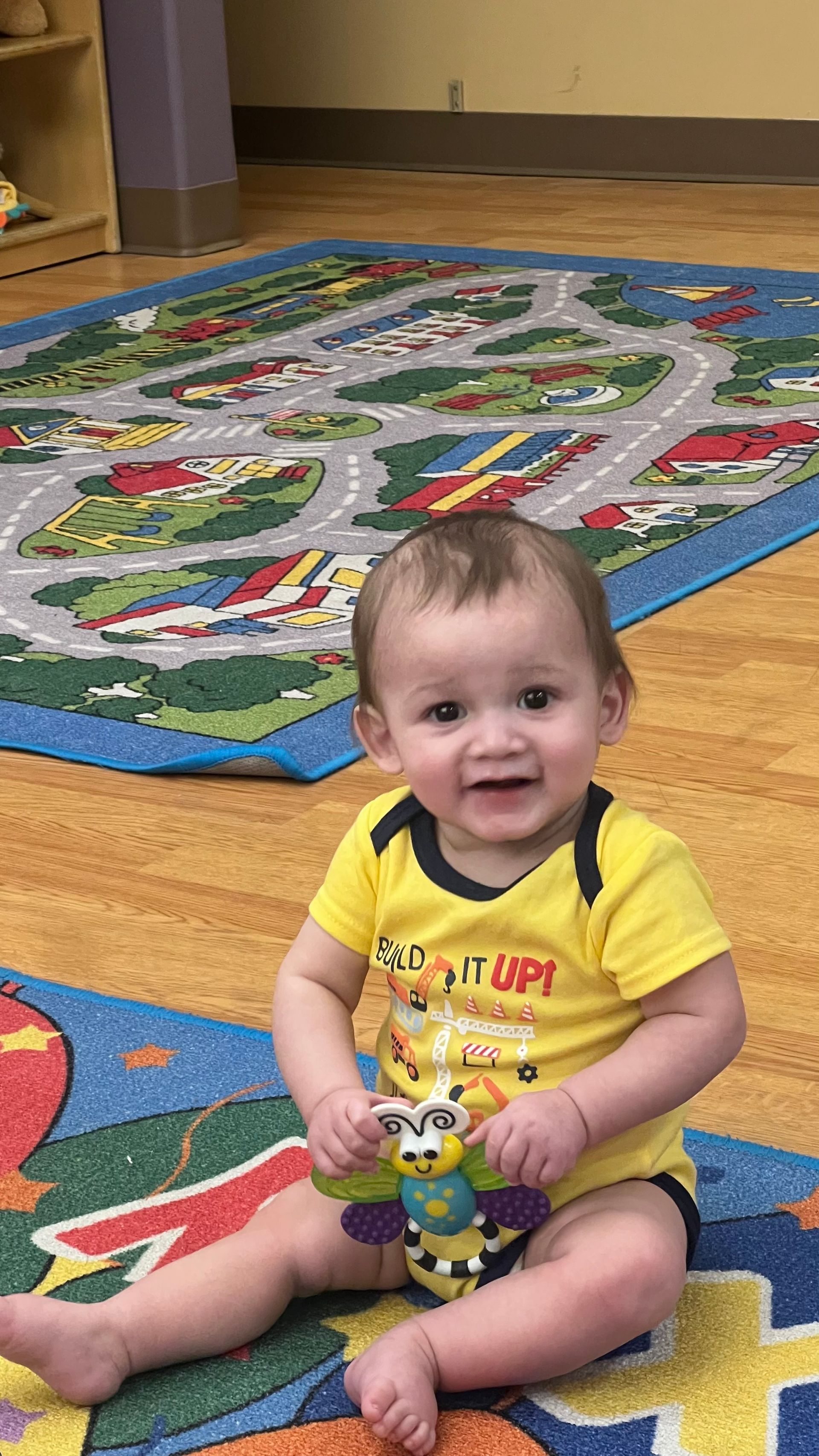 A smiling baby sits on a colorful play mat holding a small toy butterfly.